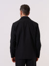 A model wears the Black Tropical Suiting Two Button Jacket over a dark top, shown from the back against a plain white background. The image emphasizes the jacket's unstructured silhouette, clean drape, and the two-piece sleeve construction. The smooth, lightweight texture of the tropical suiting fabric is visible. No mannequin is present in the image.