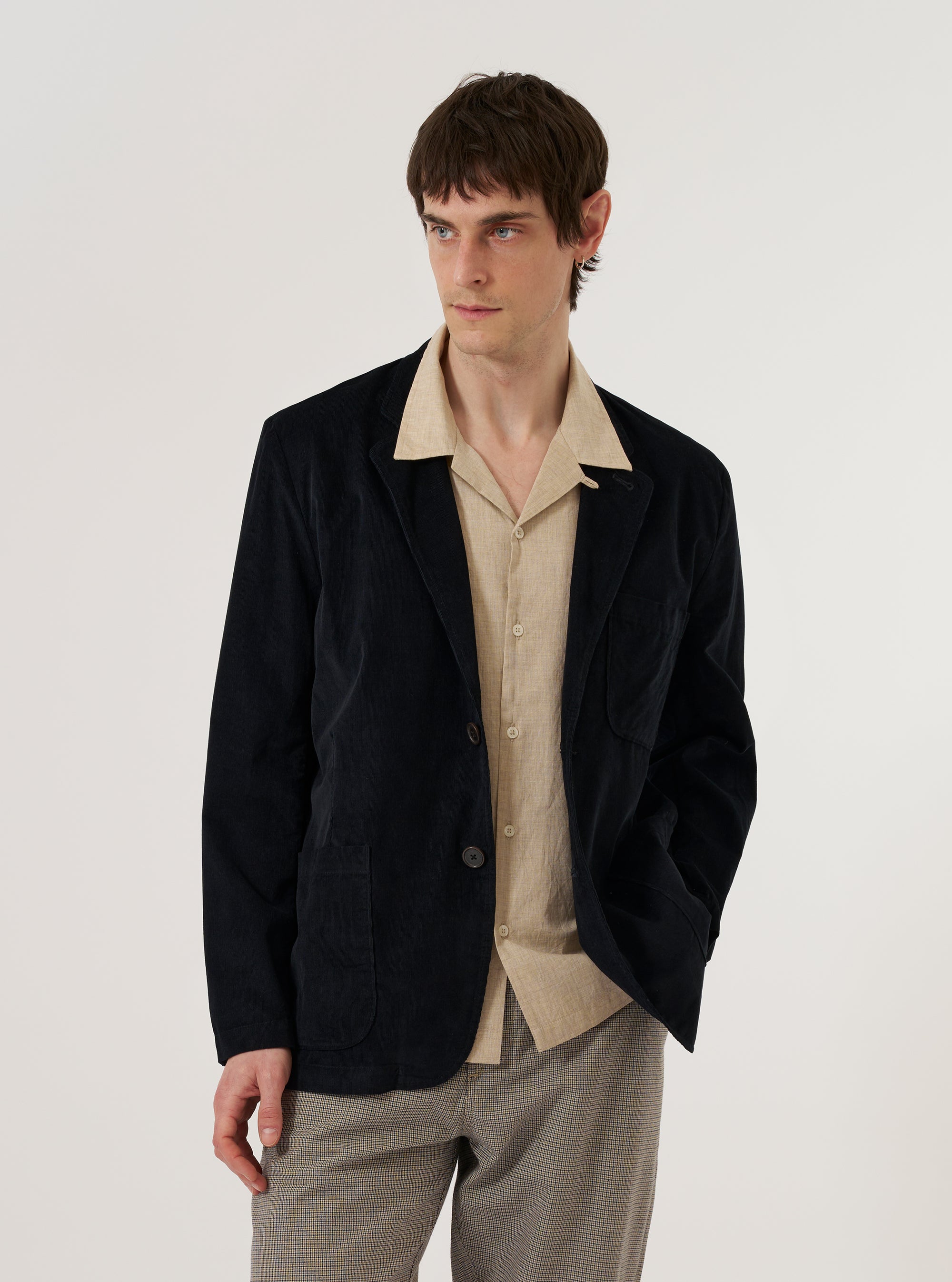 A model wears the Black Organic Fine Cord Two Button Jacket over a light-colored shirt, standing against a plain white background. The jacket features a classic two-button front closure, a notched lapel, and two large patch pockets at the waist. The fine-wale organic cotton corduroy fabric has a visible vertical texture in a deep black color.