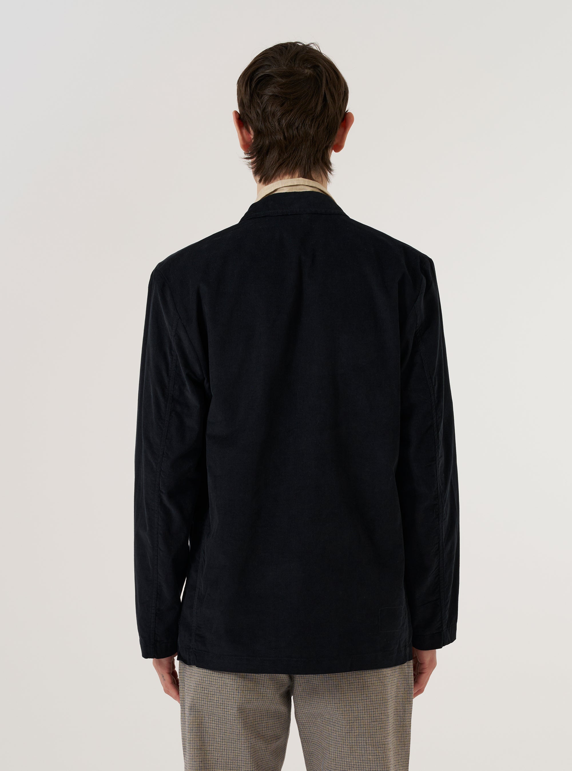 A model wears the Black Organic Fine Cord Two Button Jacket over a light-colored shirt, shown from a rear three-quarter angle against a plain white background. The jacket features a structured back with a central vent and a subtle fine-cord texture. The model is standing with their hands by their sides.