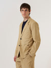 A model wears the Sand Organic Fine Cord Two Button Jacket over a light-colored shirt, shown from a rear three-quarter angle against a plain white background. The jacket features a relaxed fit, a central vent at the hem, and two-piece sleeves. The sand-colored organic fine cord fabric has a visible vertical ribbed texture.