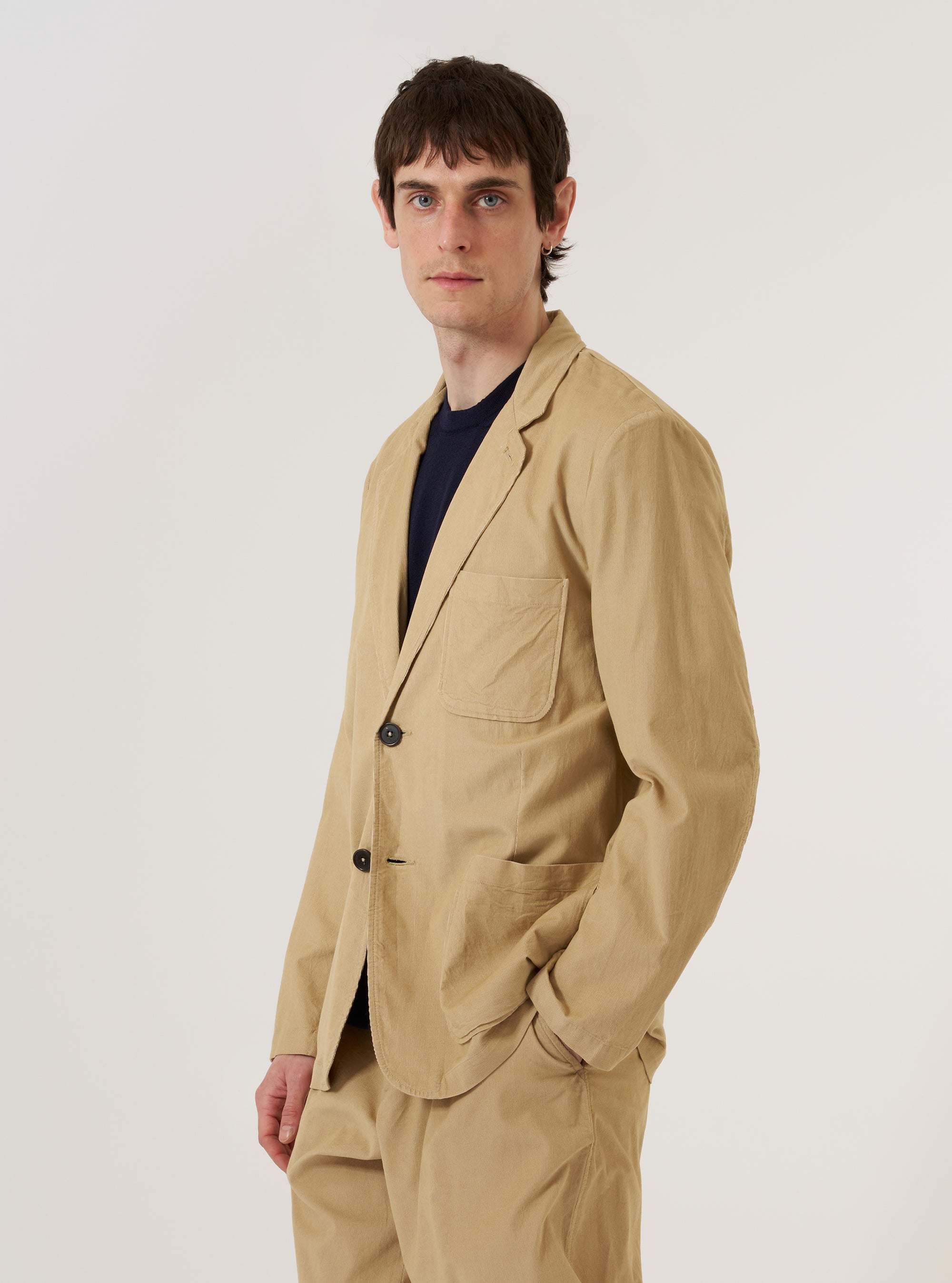 A model wears the Sand Organic Fine Cord Two Button Jacket over a light-colored shirt, shown from a rear three-quarter angle against a plain white background. The jacket features a relaxed fit, a central vent at the hem, and two-piece sleeves. The sand-colored organic fine cord fabric has a visible vertical ribbed texture.