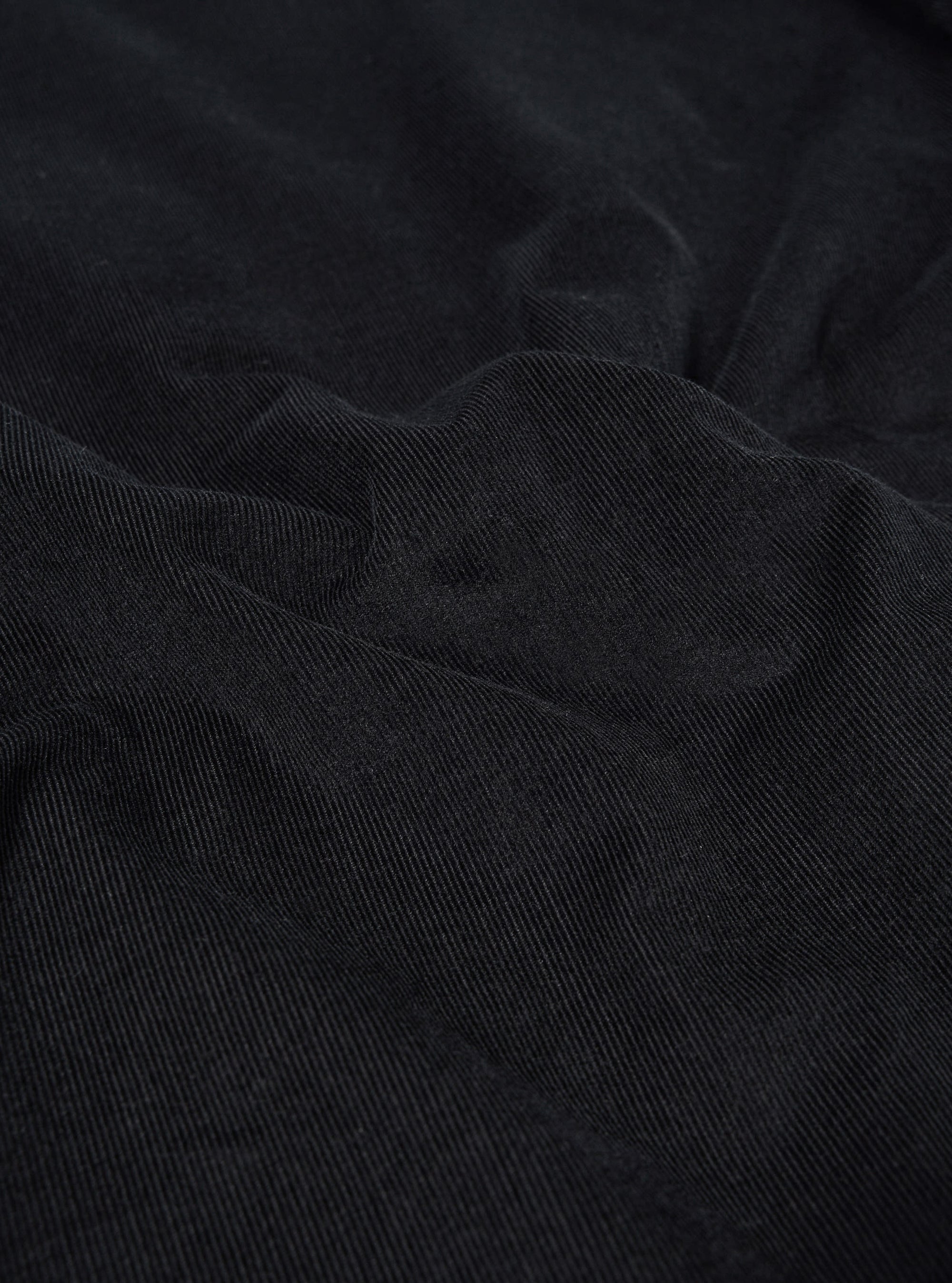 A close-up swatch of the Black Organic Fine Cord Two Button Jacket highlights the fine-wale texture and vertical ribbed pattern of the black organic cotton corduroy fabric. The material fills the frame, providing a detailed view of the garment's color and weave.