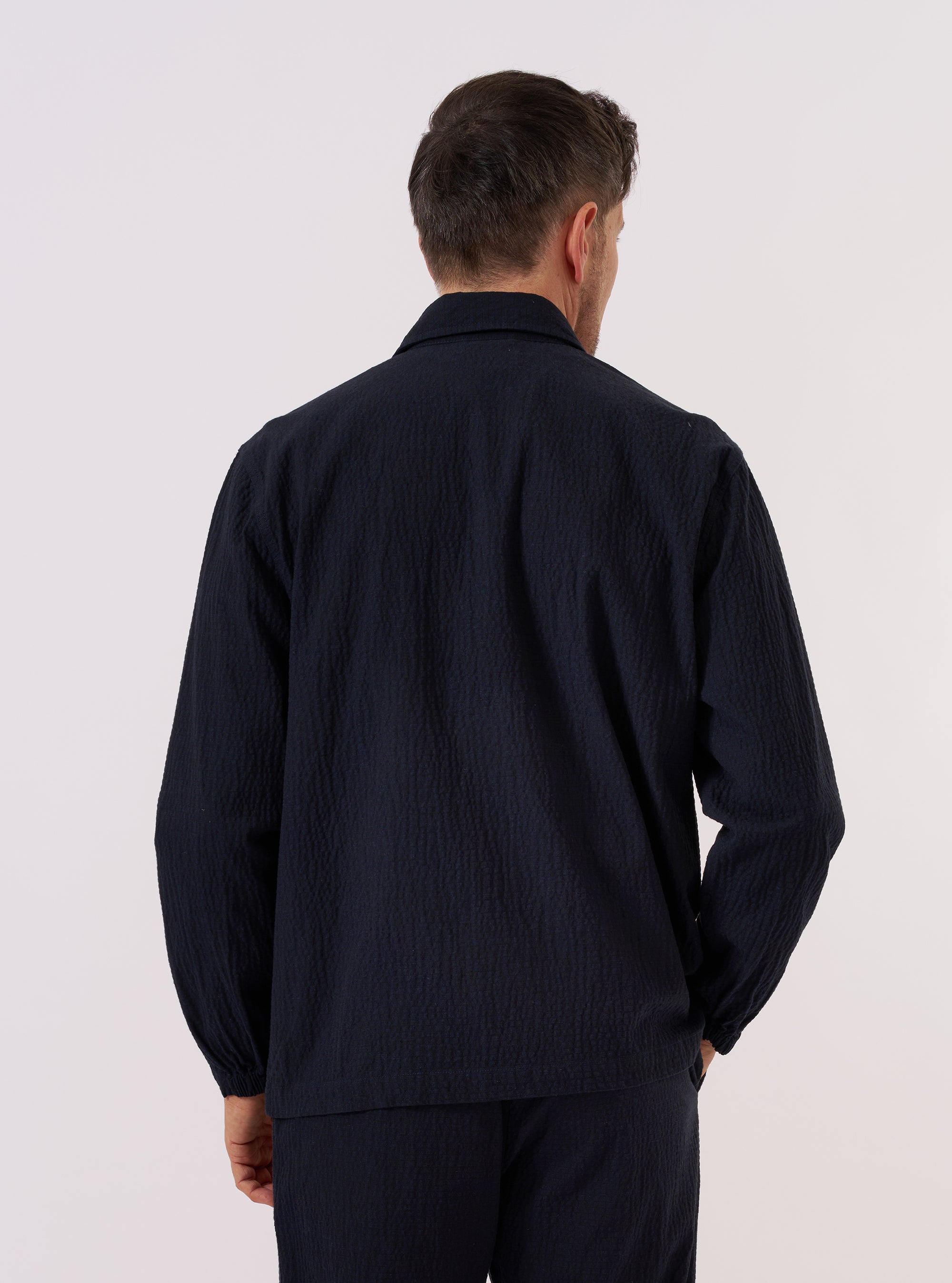 A model wears the Dark Navy Ospina Cotton Two Button Jacket, shown from the back against a plain white background. The jacket features a clean back panel with a central rear vent and a straight hem. The dark navy fabric highlights a distinct puckered seersucker texture throughout.