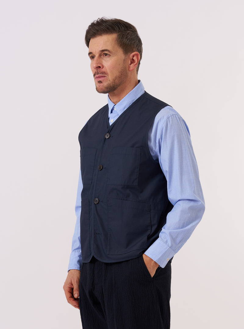A model wears the Navy Recycled Polytech City Waistcoat over a light-colored shirt. The waistcoat features a button-down front, a V-neckline, and three prominent patch pockets on the front. The image is a medium shot captured against a plain, light grey background.