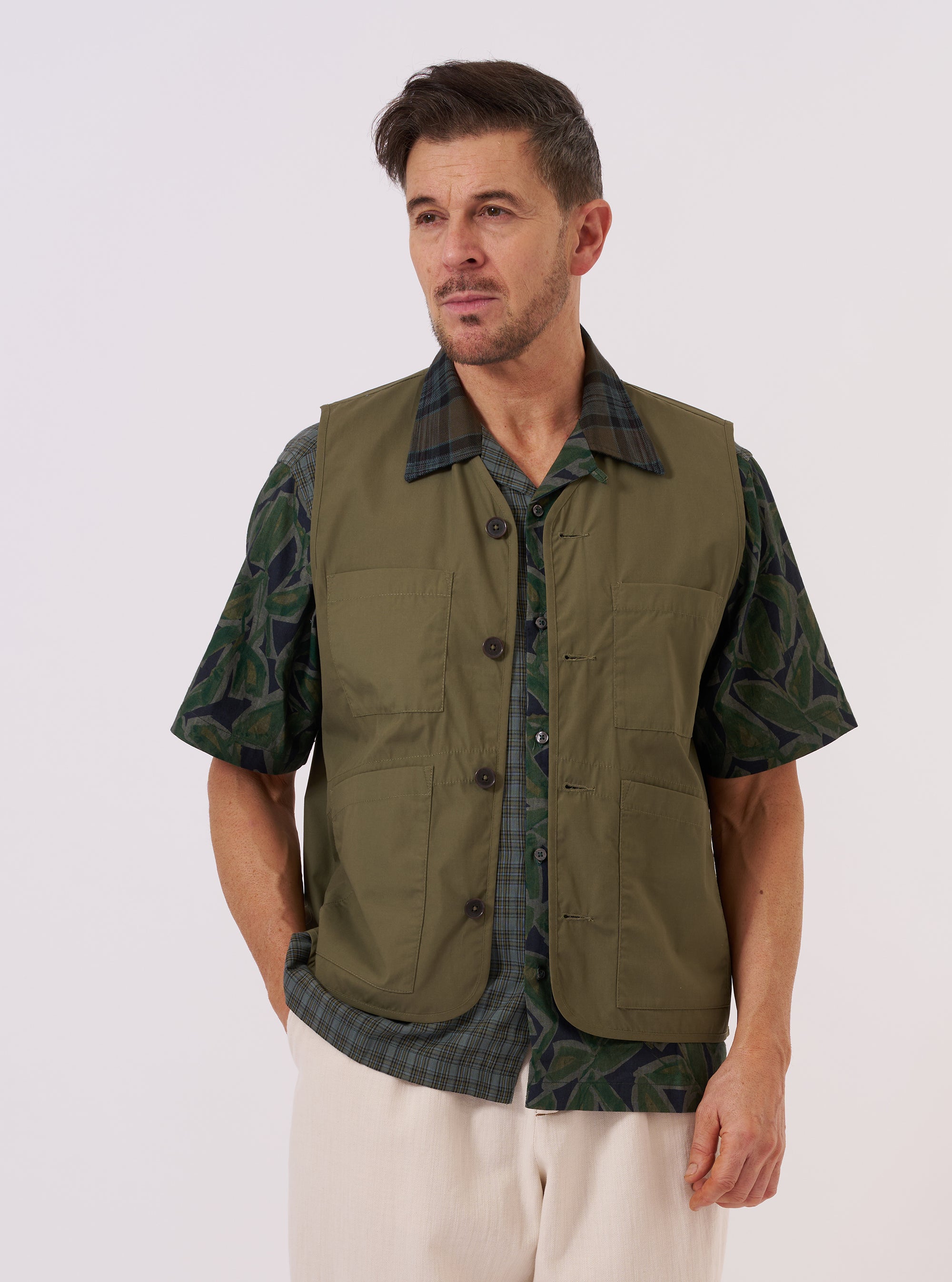 A model wears the Olive Recycled Polytech City Waistcoat against a plain white background. The model is wearing the waistcoat over a light-colored long-sleeved shirt and dark trousers. The garment features a V-neckline, a five-button front closure, and two large patch pockets at the waist. The olive green fabric has a smooth, technical finish.