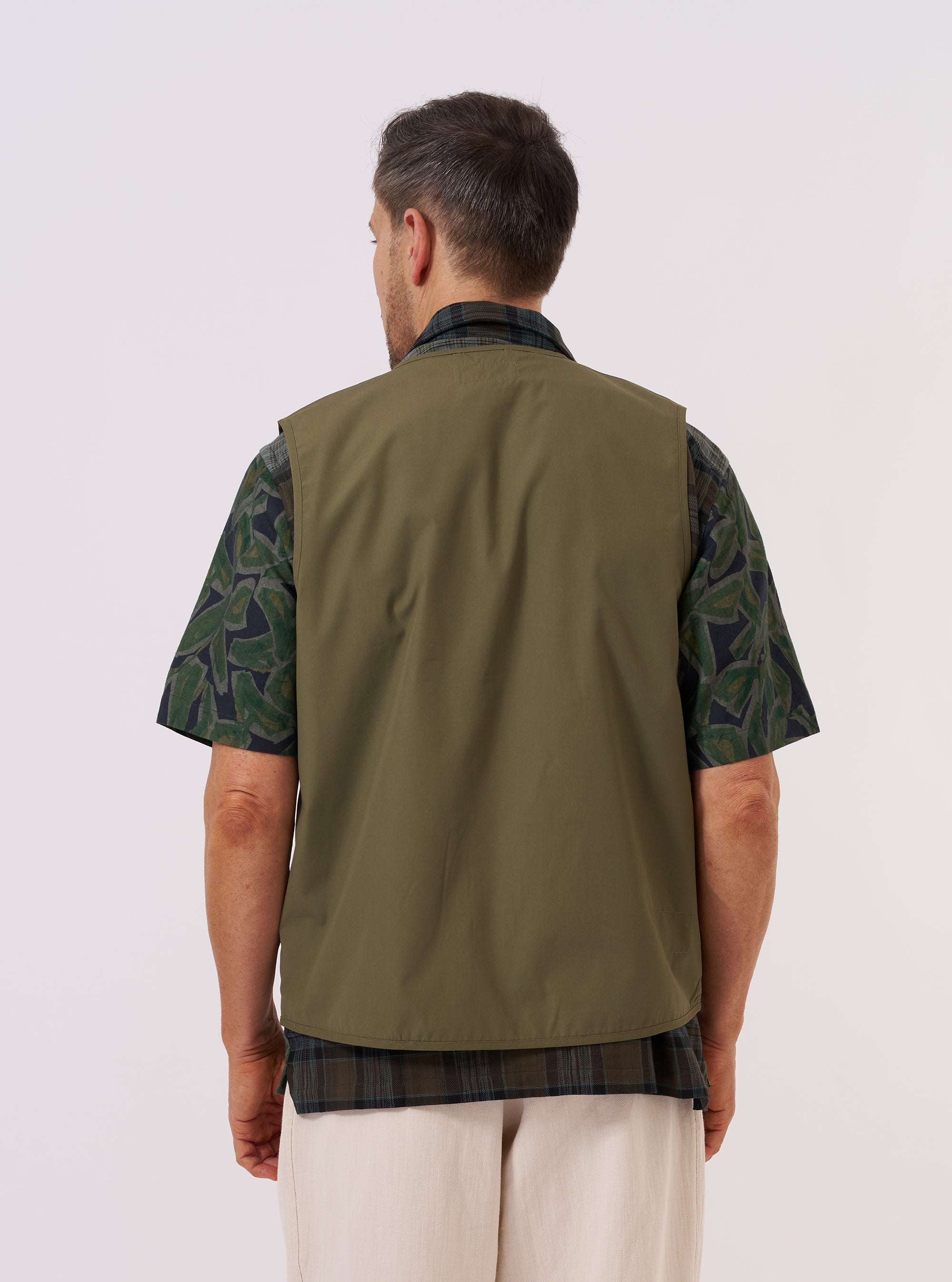 A model is wearing the Olive Recycled Polytech City Waistcoat over a white long-sleeved shirt against a plain white background. This side-profile view highlights the garment's clean lines, sleeveless design, and the smooth finish of the olive green fabric. The waistcoat is shown buttoned up, featuring a structured collar and a straight hem.