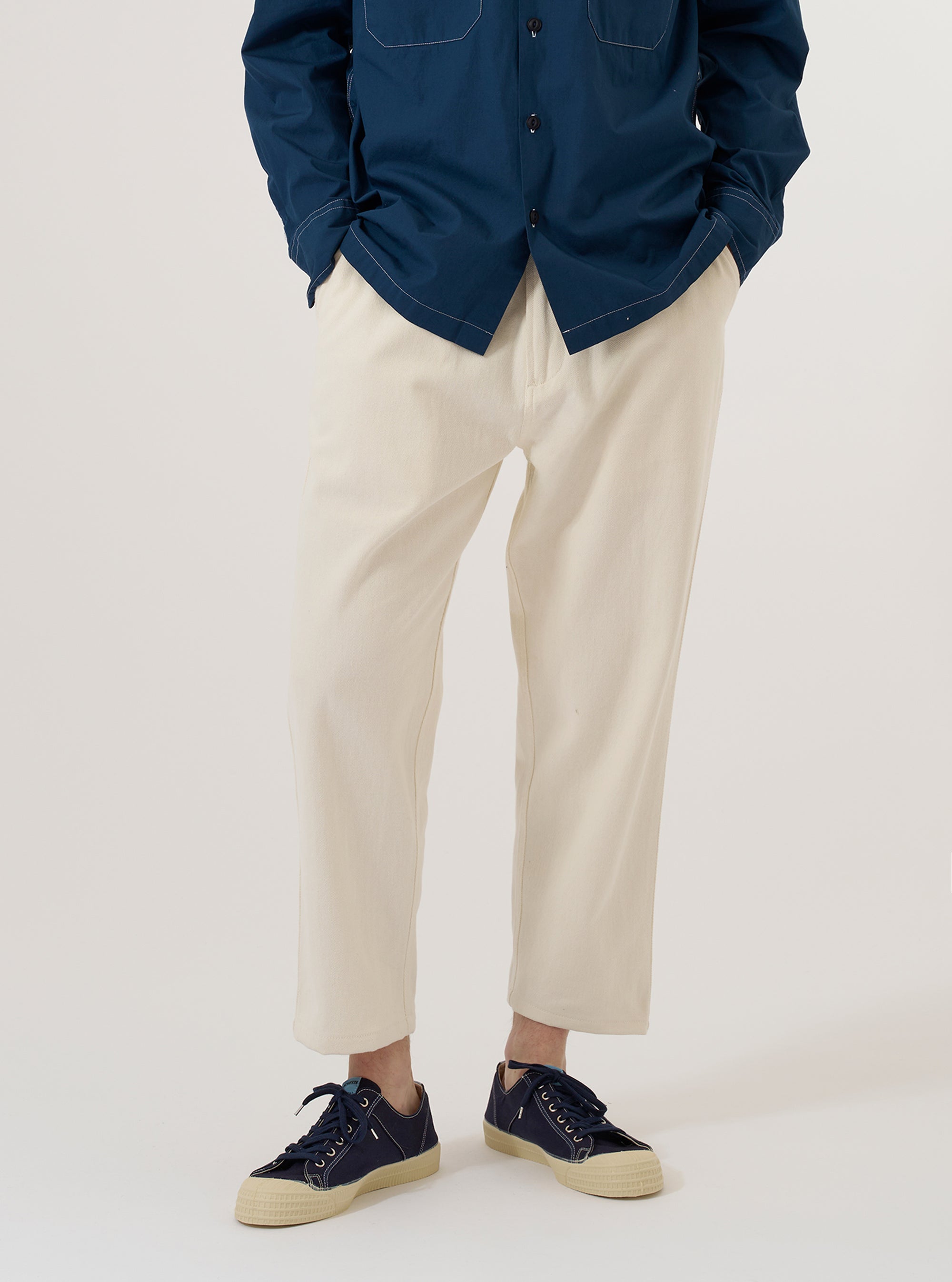 A model wears the Ecru Slub Drill Hi Water Trouser against a plain, neutral background. The trousers feature a slim, cropped fit and a drawstring waistband. The ecru-colored cotton fabric has a visible slub drill texture. The model is shown from the waist down, highlighting the garment's length and structured design.
