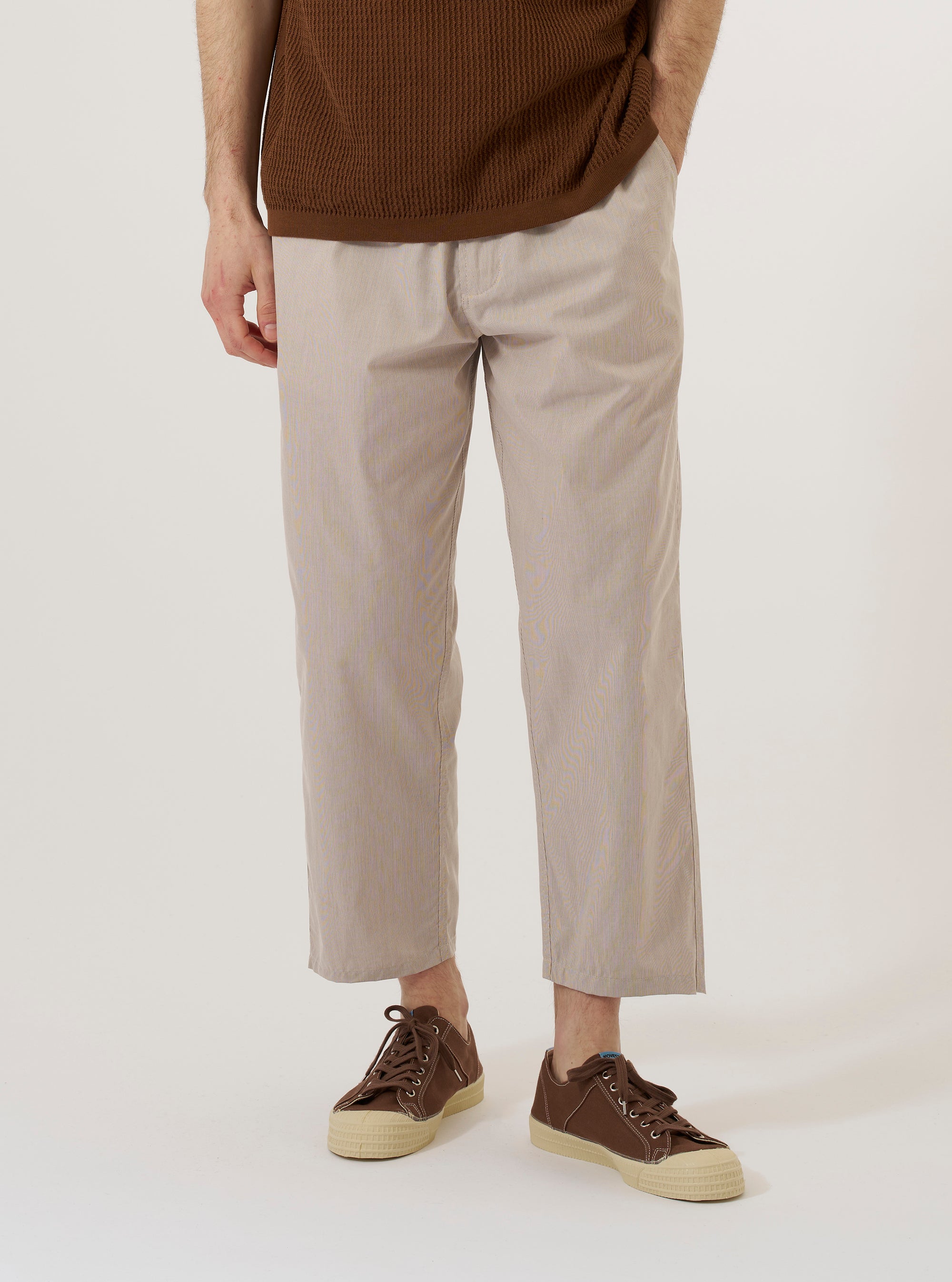 A model wears the Brown Kennedy Stripe Hi Water Trouser, shown from the waist down against a plain white background. The trousers feature a vertical pinstripe pattern, a relaxed fit with a tapered leg, and a slightly cropped length that hits above the ankle. They are designed with a drawstring waist and slanted side pockets.
