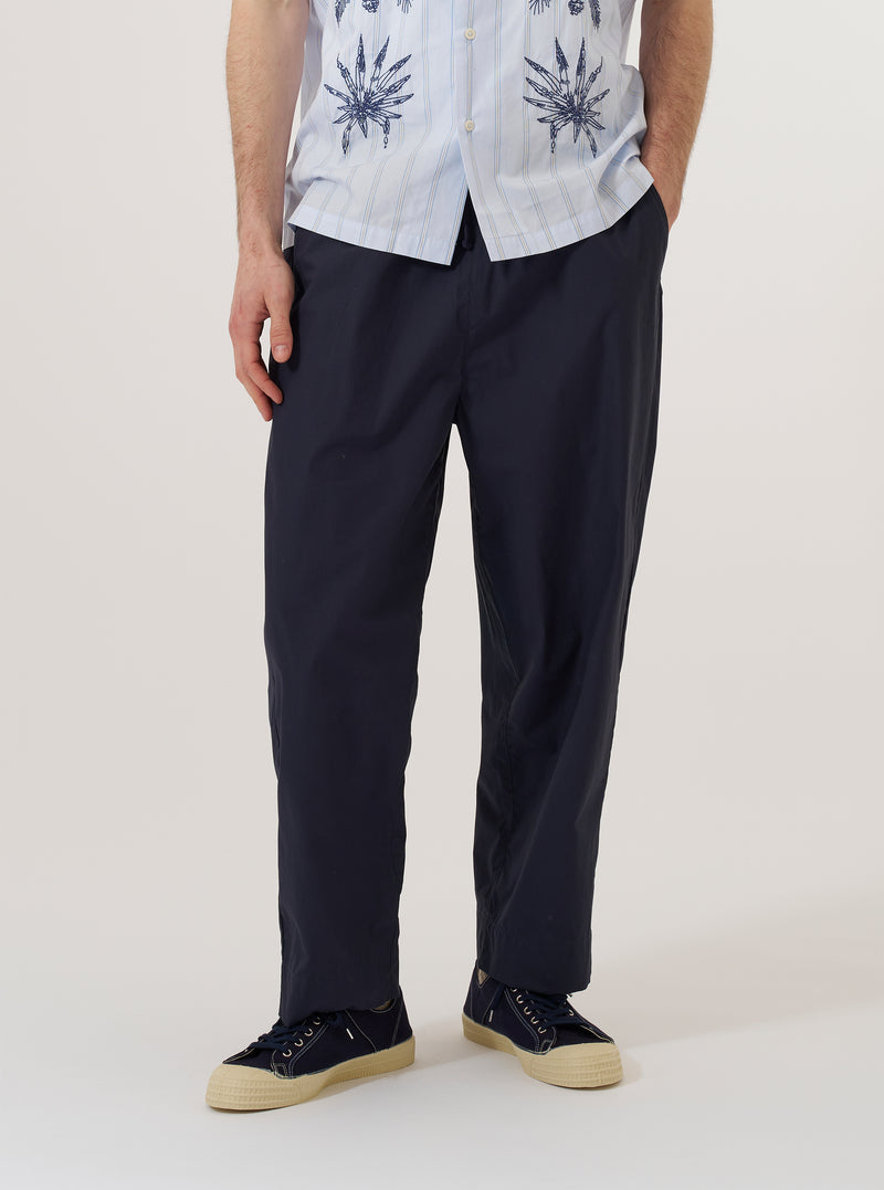 A model wears the Navy Peached Poplin Judo Pant, shown from the waist down against a plain white background. The image highlights the trousers' simple, straight-leg silhouette and relaxed fit. The model is wearing the navy blue cotton pants with the elasticated waistband visible, and the fabric has a soft, matte peached finish. One front pocket is visible on the side.