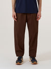 A model wears the Brown Peached Poplin Judo Pant against a plain white background. The image shows a front view of the trousers, highlighting the relaxed fit, elasticated drawstring waistband, and the smooth texture of the brown cotton poplin fabric. The model is wearing a light-colored top tucked into the pants, which feature a slightly tapered leg.