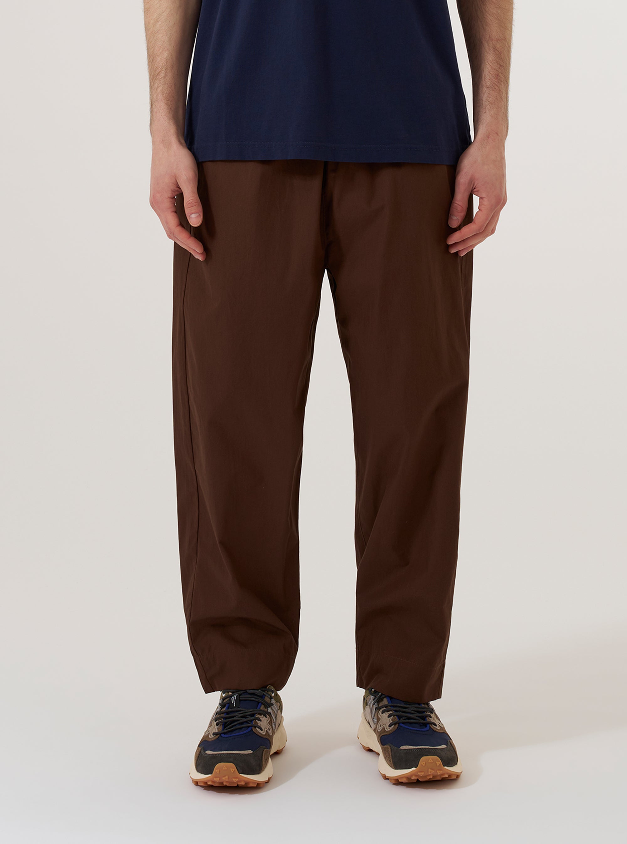 A model wears the Brown Peached Poplin Judo Pant against a plain white background. The image shows a front view of the trousers, highlighting the relaxed fit, elasticated drawstring waistband, and the smooth texture of the brown cotton poplin fabric. The model is wearing a light-colored top tucked into the pants, which feature a slightly tapered leg.