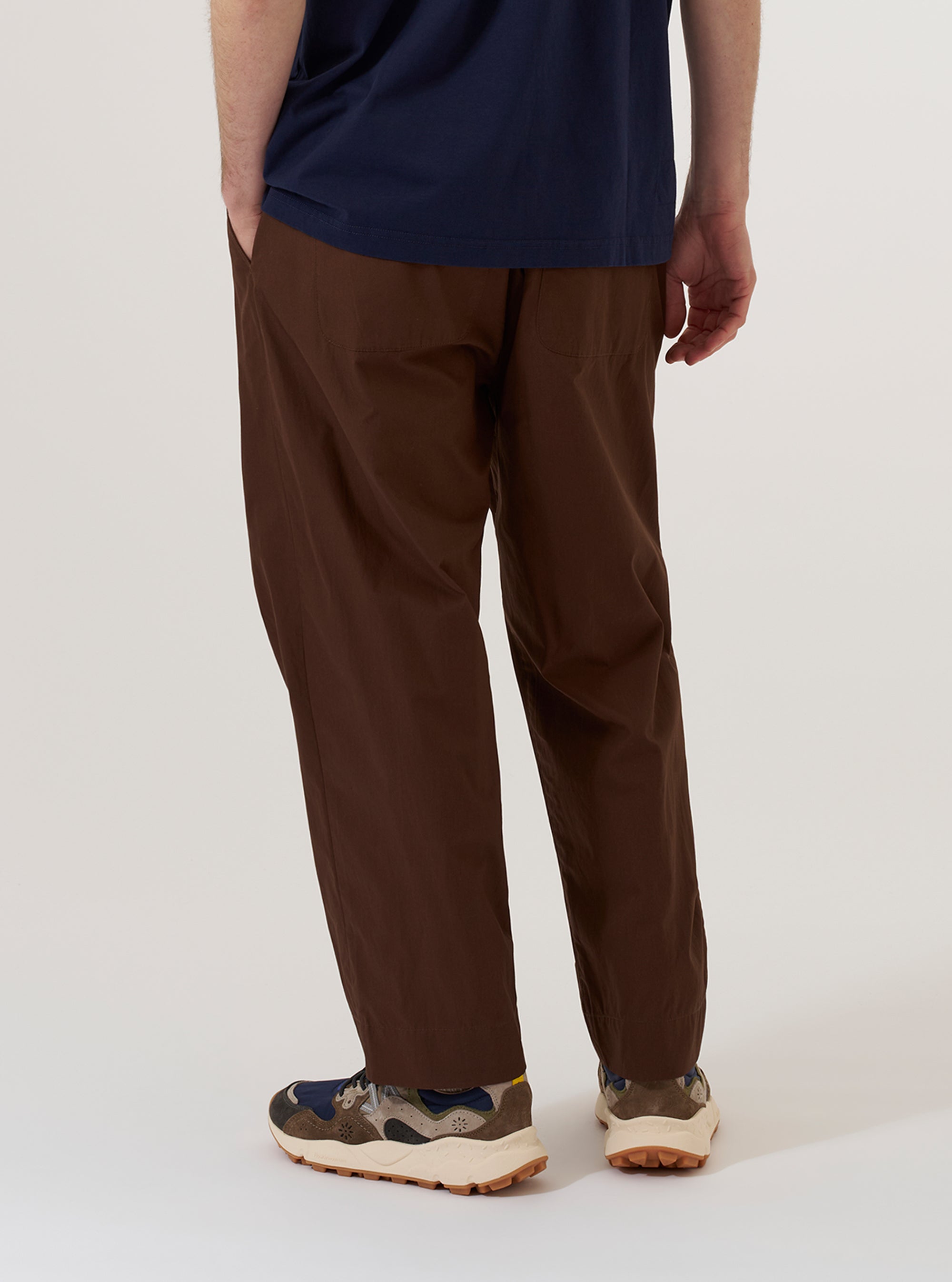 A model wears the Brown Peached Poplin Judo Pant, shown from the waist down against a plain white background. The pants feature a loose, relaxed fit with a straight leg and are made from a smooth brown cotton poplin. The model is shown from a rear three-quarter angle, highlighting the clean construction and the way the fabric drapes. No mannequin is present in the image.