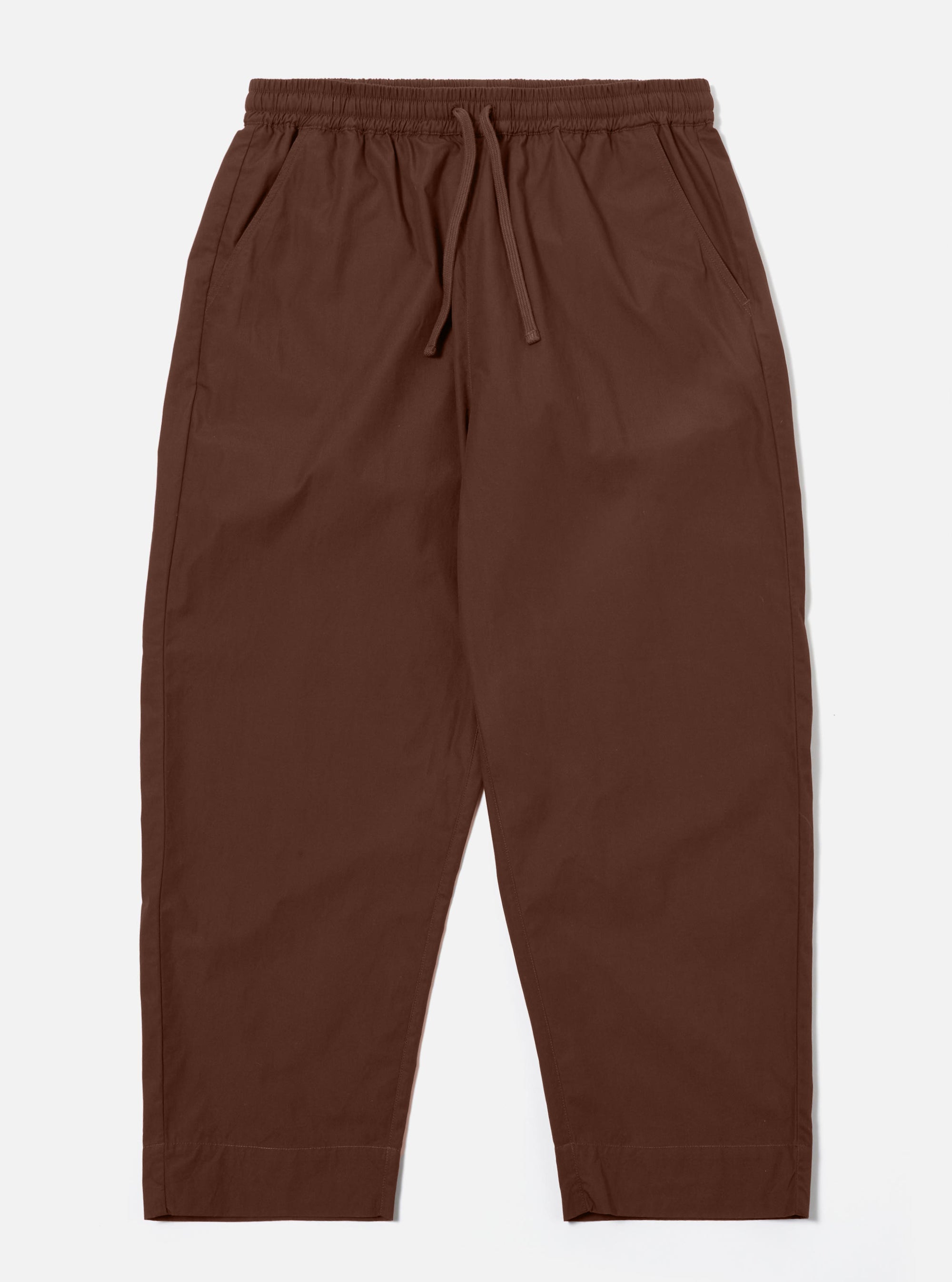 The Brown Peached Poplin Judo Pant is shown as a flat lay against a white background. The trousers feature a relaxed, straight-leg silhouette with an elasticated waistband and an integrated drawstring. Two front pockets and a subtle hanging loop on the waistband are visible. The brown cotton fabric has a soft, matte finish characteristic of peached poplin.