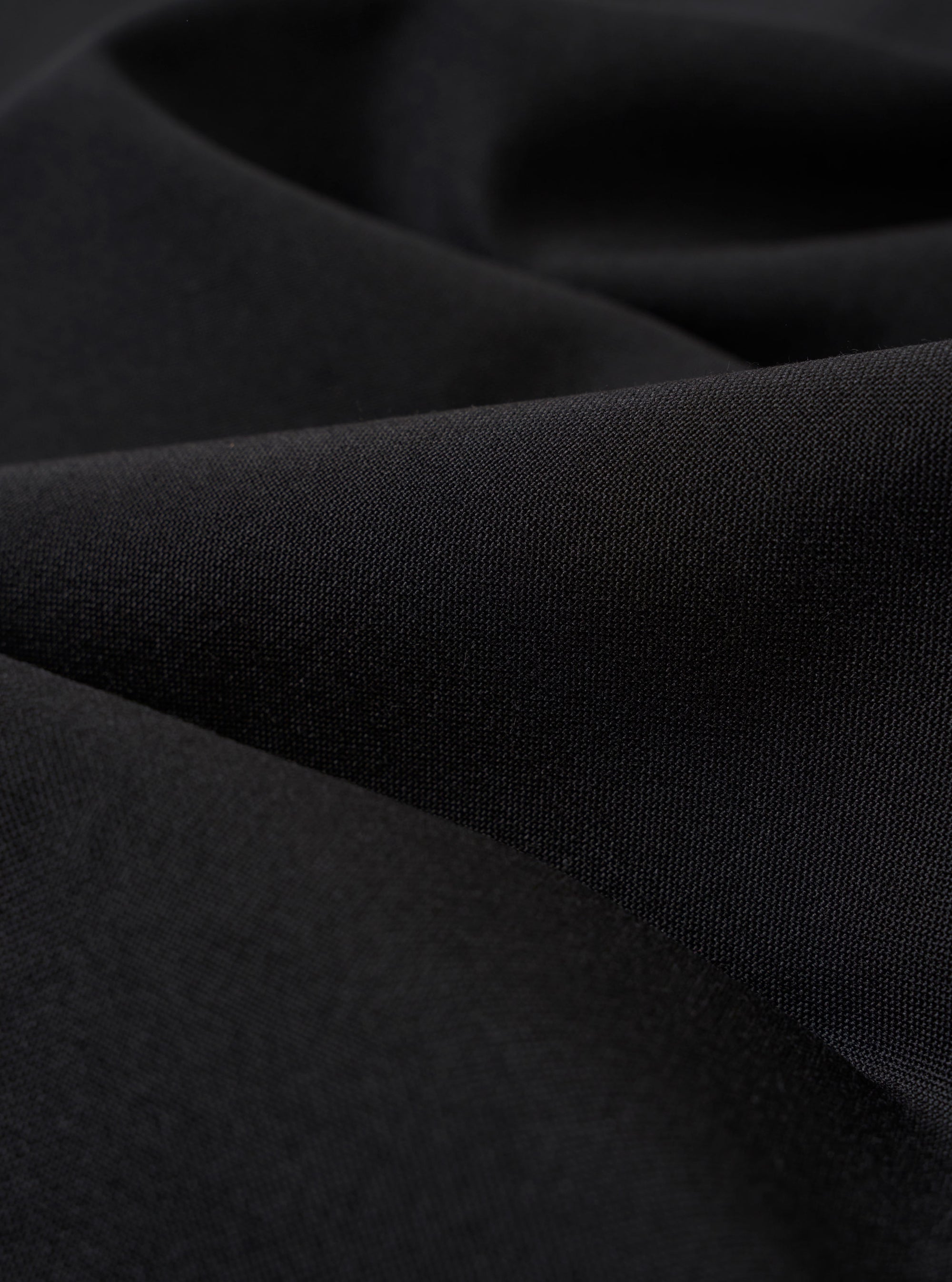 A close-up fabric swatch of the Black Tropical Suiting Military Chino highlights the smooth, technical texture and deep black color of the polyester and wool blend material. The fabric fills the entire frame, showcasing the fine weave of the tropical suiting.