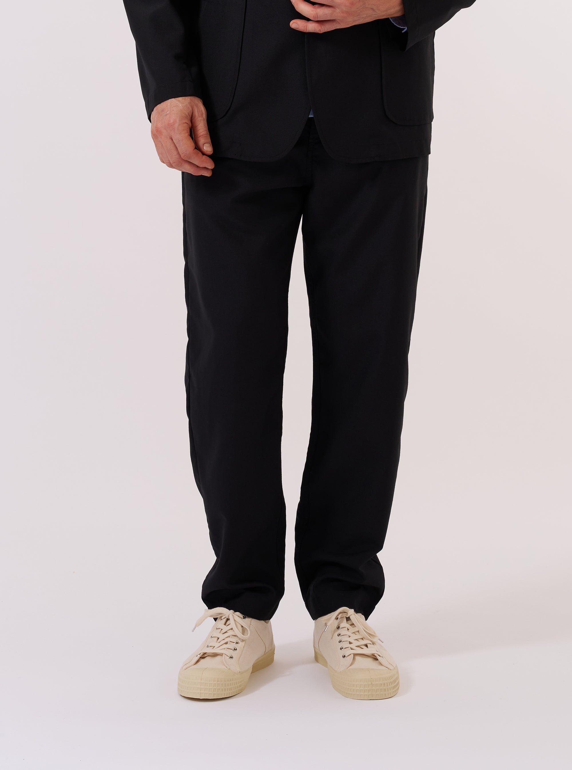 A model wears the Black Tropical Suiting Military Chino, shown from the waist down against a plain, neutral background. These black trousers feature a slim-straight fit that tapers towards the ankle, with visible front pockets and a mid-rise waist. The model is standing with one leg slightly forward, highlighting the clean drape of the lightweight polyester-wool blend fabric. No mannequin is present in the image.