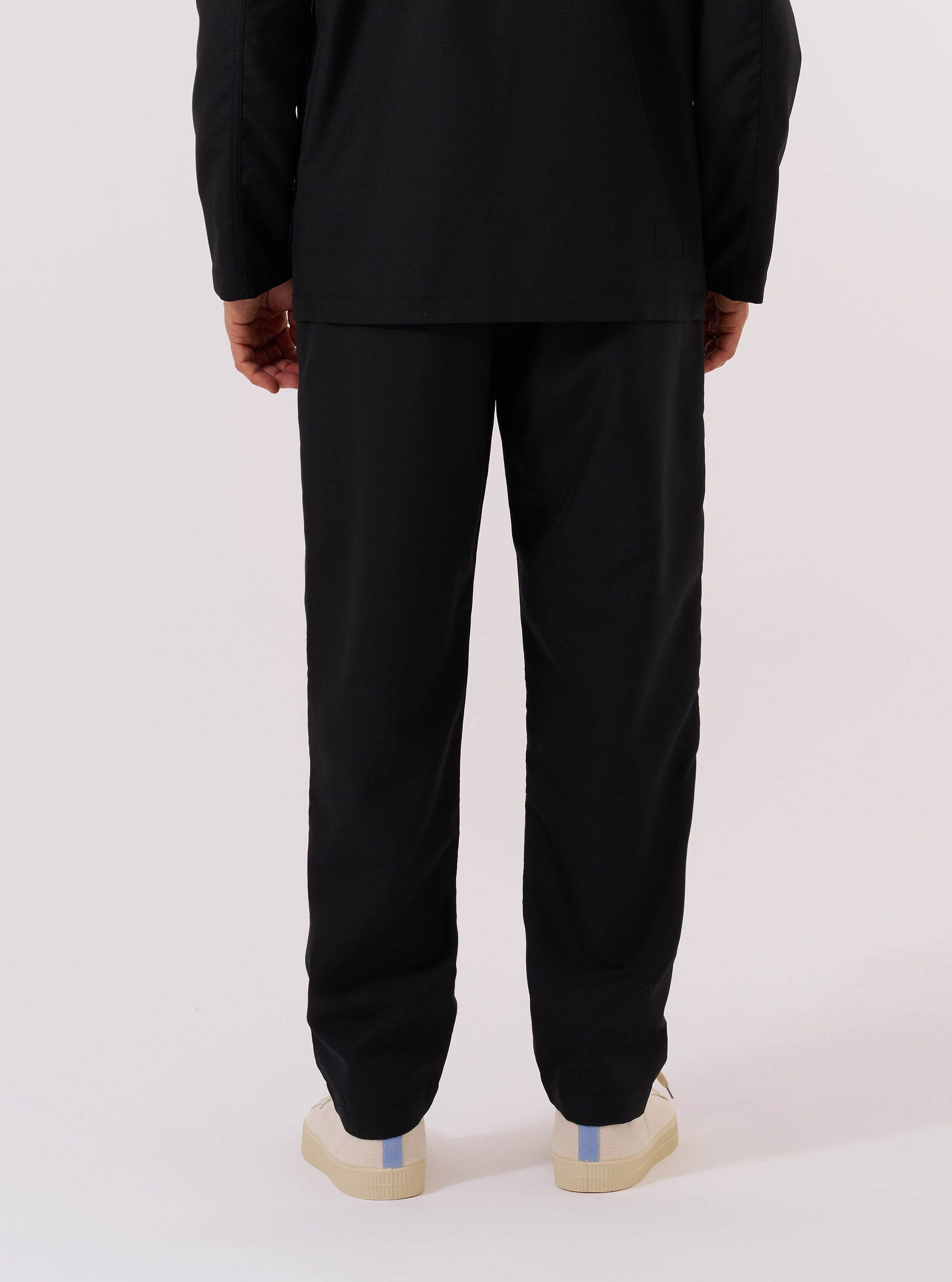 A model is wearing the Black Tropical Suiting Military Chino against a plain white background. This rear view highlights the trousers' tapered leg, two jetted back pockets with button closures, and the smooth, lightweight texture of the black polyester and wool blend fabric. The model is shown from the waist down wearing dark socks and shoes. No mannequin is present in the image.