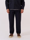 A model wears the Navy Tropical Suiting Military Chino against a plain white background. The image shows a front view of the trousers, highlighting the mid-rise waist, button fly, and two front pockets. The lightweight navy fabric has a clean, smooth appearance, and the leg shows a gradual taper towards the ankle. The model is wearing a light-colored shirt tucked into the chinos and white socks with loafers.