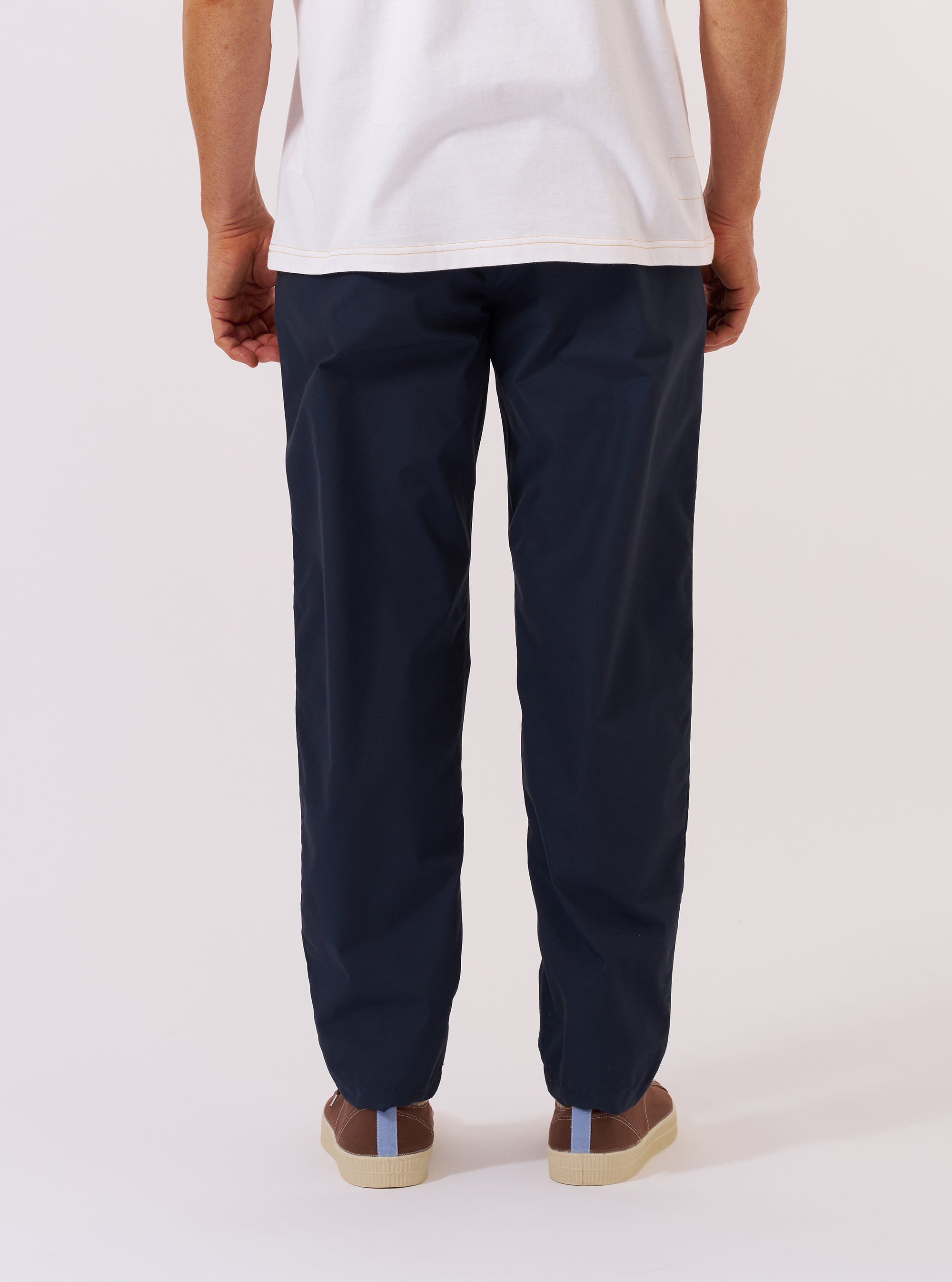 A model wears the Navy Recycled Polytech Military Chino, shown from the rear three-quarter angle against a plain white background. The image highlights the tapered leg, the clean finish of the navy recycled polytech fabric, and a rear jetted buttoned pocket. The model is also wearing white socks and brown shoes.