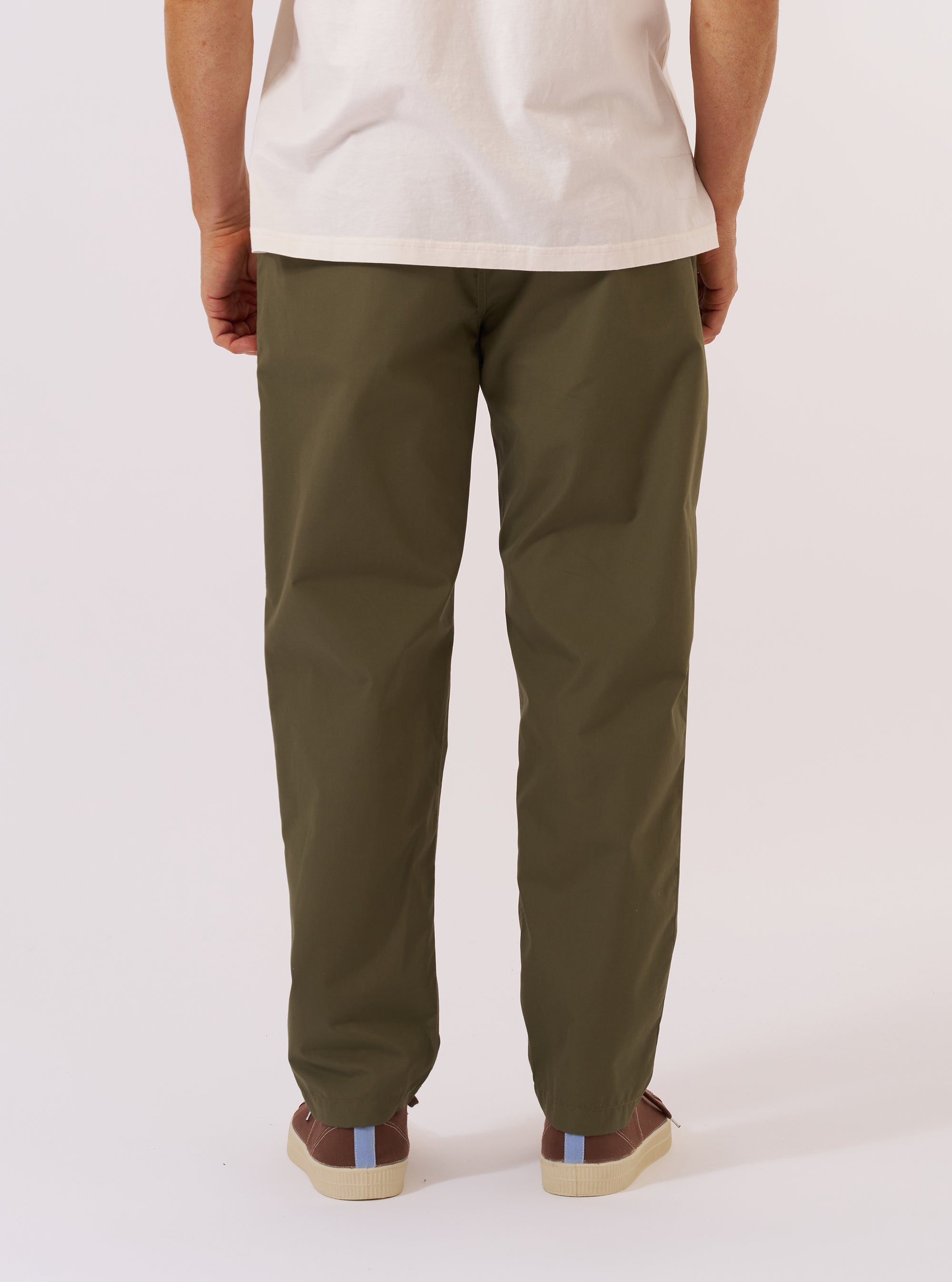 A model wears the Olive Recycled Polytech Military Chino, shown from the waist down against a plain, neutral studio background. The image highlights the wide but tapered fit of the trousers, which are made from a smooth olive green recycled polyester and cotton blend. The model is captured from a side angle, showcasing the tailored silhouette and the clean finish of the technical fabric. No mannequin is present in the image.