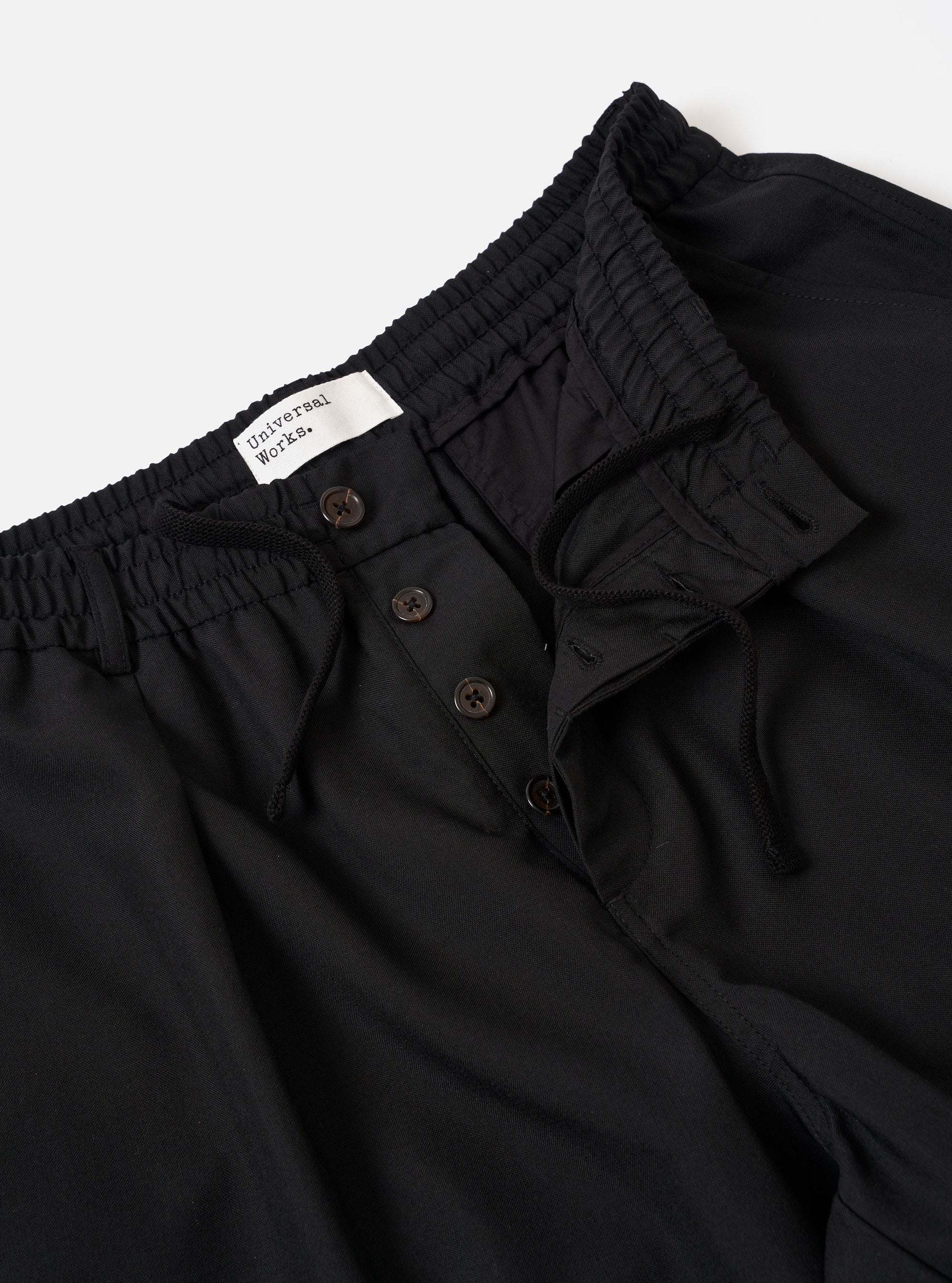 The Black Tropical Suiting Pleated Track Pant is shown from the back as a flat lay against a plain white background. The image highlights the two rear pockets, including one with a button-down flap, and the elasticated waistband. The lightweight black tropical suiting fabric, made of a polyester and wool blend, appears smooth and crisp.