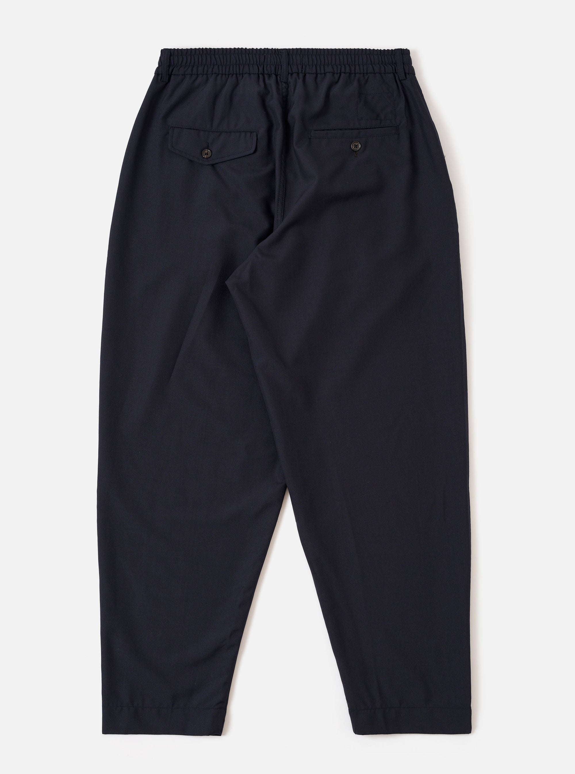 The Navy Tropical Suiting Pleated Track Pant is shown from the back as a flat lay against a plain white background. The image highlights the elasticated waistband and the two rear pockets, one of which features a buttoned flap. The navy wool-blend fabric has a smooth, clean finish.