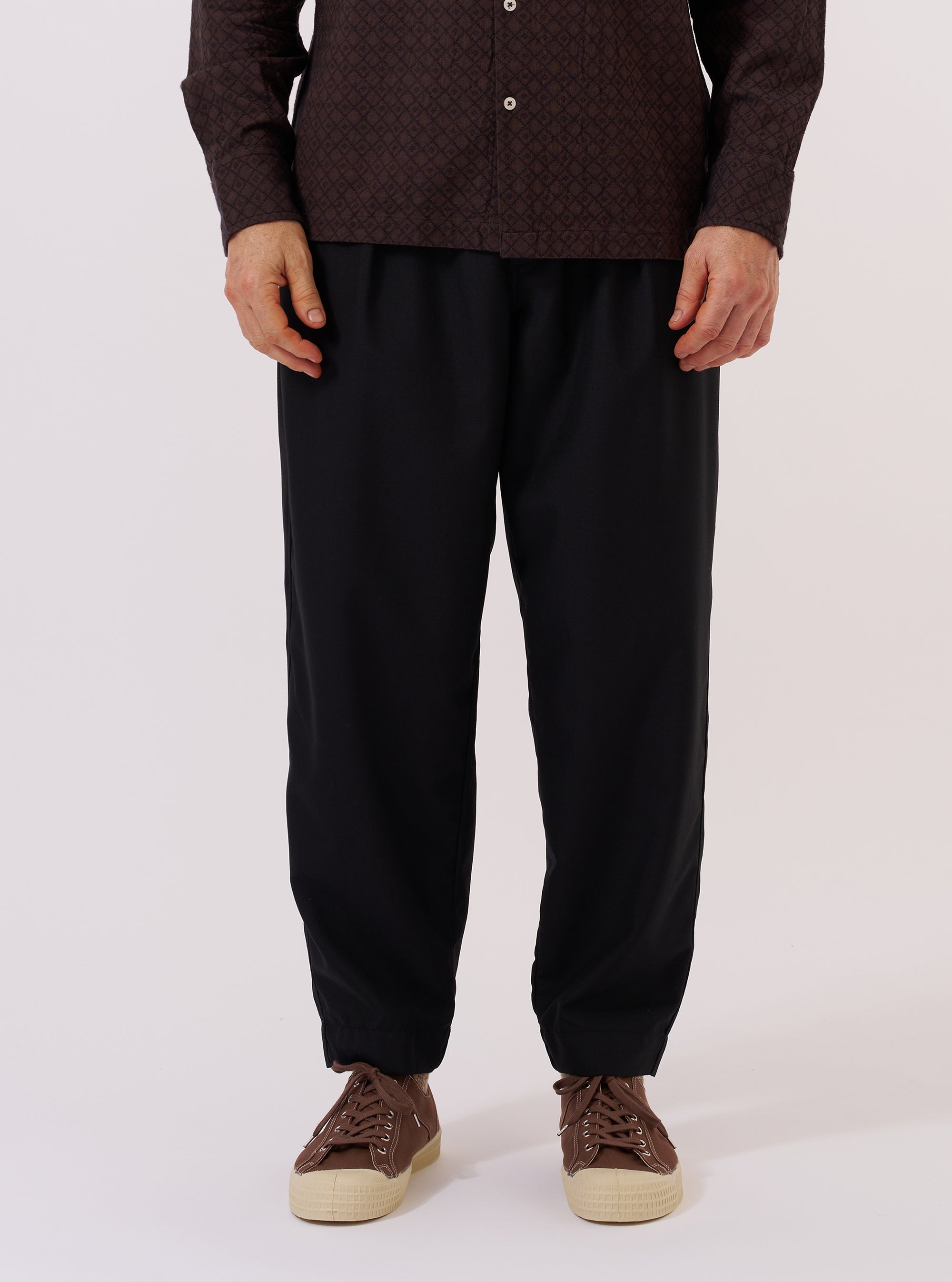 A model wears the Black Tropical Suiting Pleated Track Pant against a plain, neutral background. The trousers feature a relaxed, tapered fit with prominent front pleats and an elasticated waistband with a drawstring. The garment is made from a smooth black tropical suiting fabric and is shown from the waist down to highlight the construction and drape.