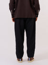 A model is wearing the Black Tropical Suiting Pleated Track Pant, shown from a rear three-quarter angle against a plain white background. The image highlights the relaxed fit and the clean, smooth texture of the black tropical suiting fabric. The trousers feature an elasticated waistband and a distinct taper toward the hem.