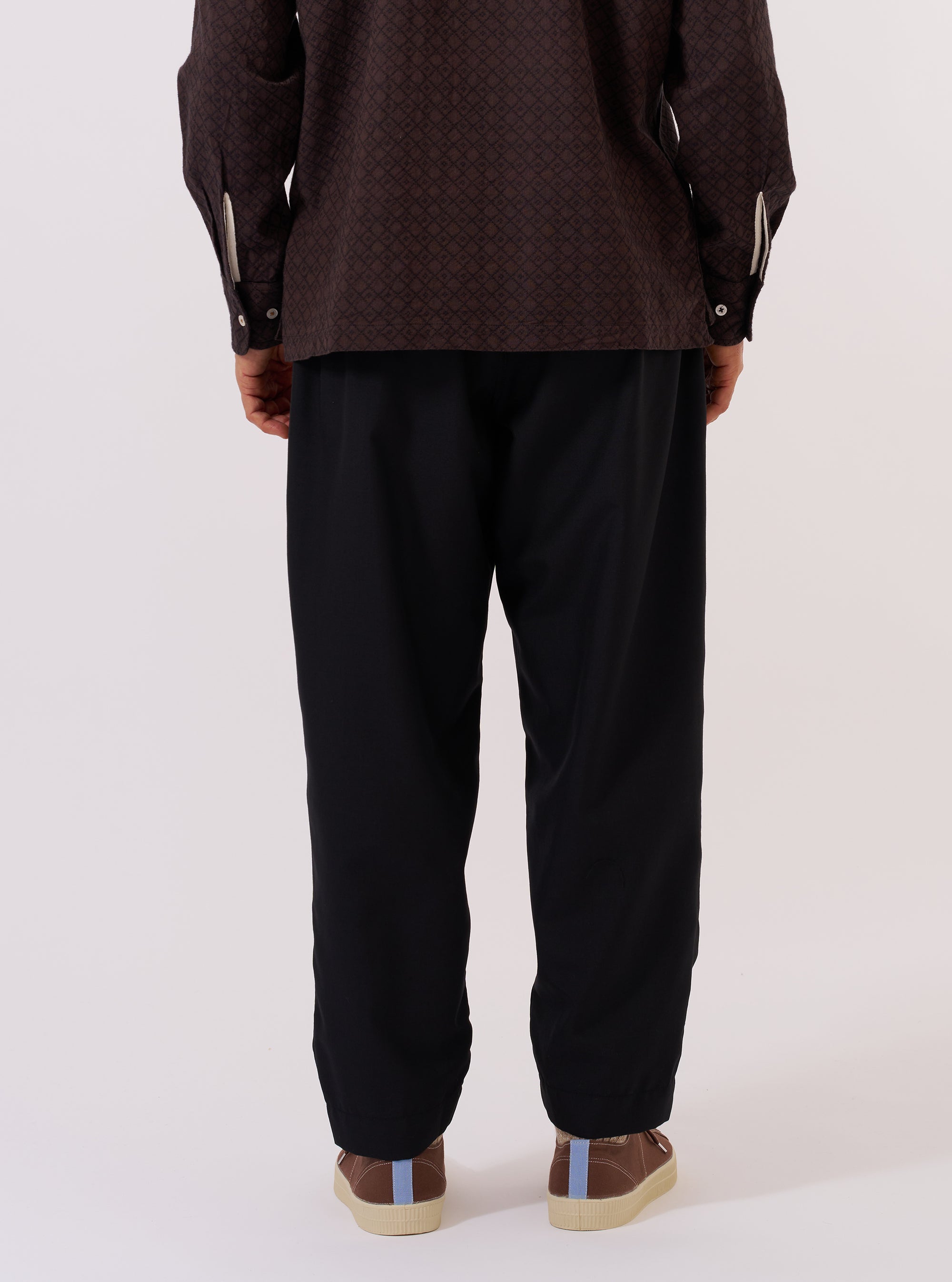 A model is wearing the Black Tropical Suiting Pleated Track Pant, shown from a rear three-quarter angle against a plain white background. The image highlights the relaxed fit and the clean, smooth texture of the black tropical suiting fabric. The trousers feature an elasticated waistband and a distinct taper toward the hem.