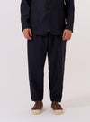 The Navy Tropical Suiting Pleated Track Pant is worn by a model and shown from the waist down against a plain white background. The image highlights the loose, tapered fit of the trousers, the single pleat at the front, and the elasticated waistband with a visible drawstring. The navy blue fabric appears smooth with a clean, minimal finish. The model is also wearing white socks and brown shoes.