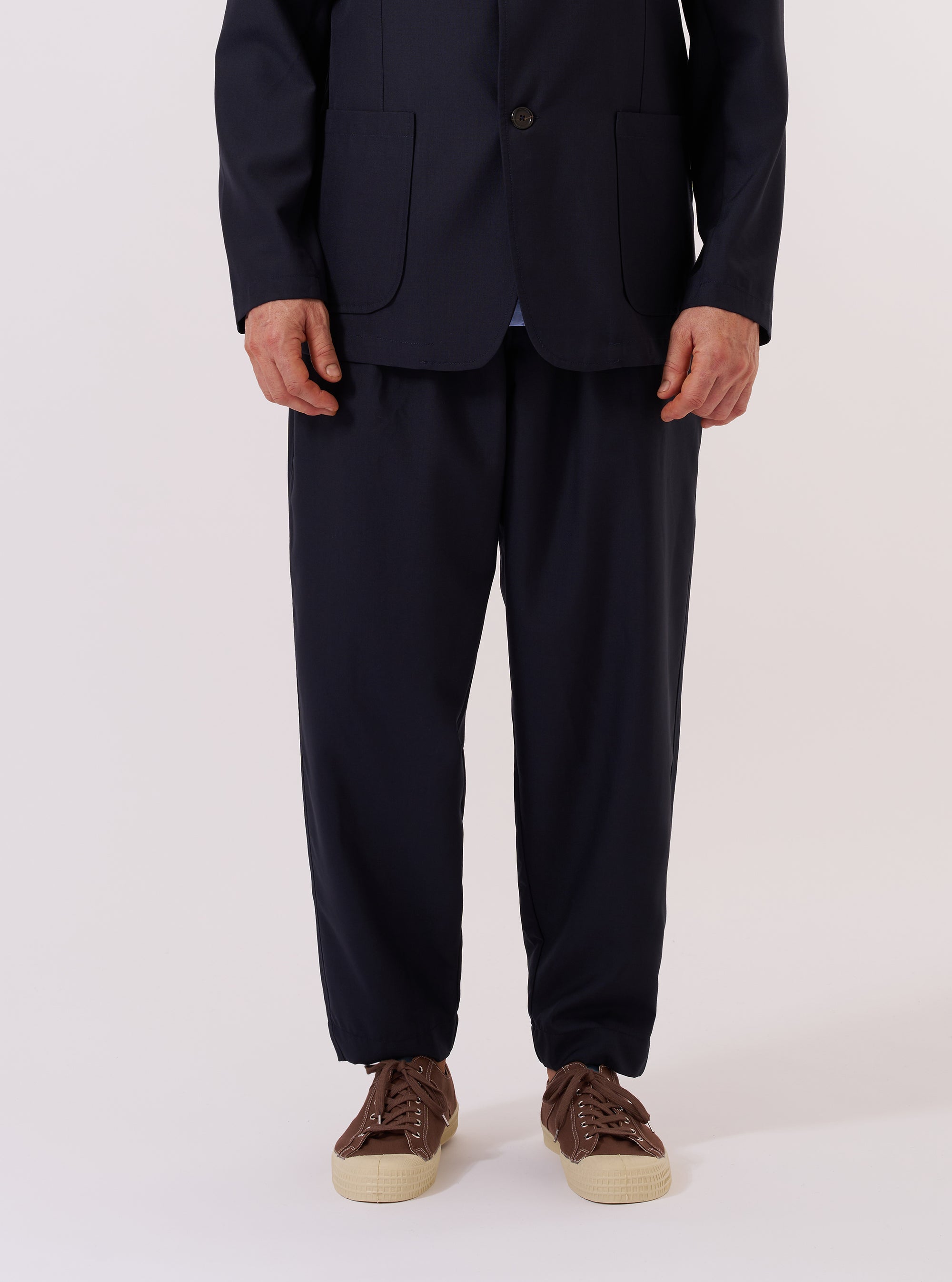 The Navy Tropical Suiting Pleated Track Pant is worn by a model and shown from the waist down against a plain white background. The image highlights the loose, tapered fit of the trousers, the single pleat at the front, and the elasticated waistband with a visible drawstring. The navy blue fabric appears smooth with a clean, minimal finish. The model is also wearing white socks and brown shoes.