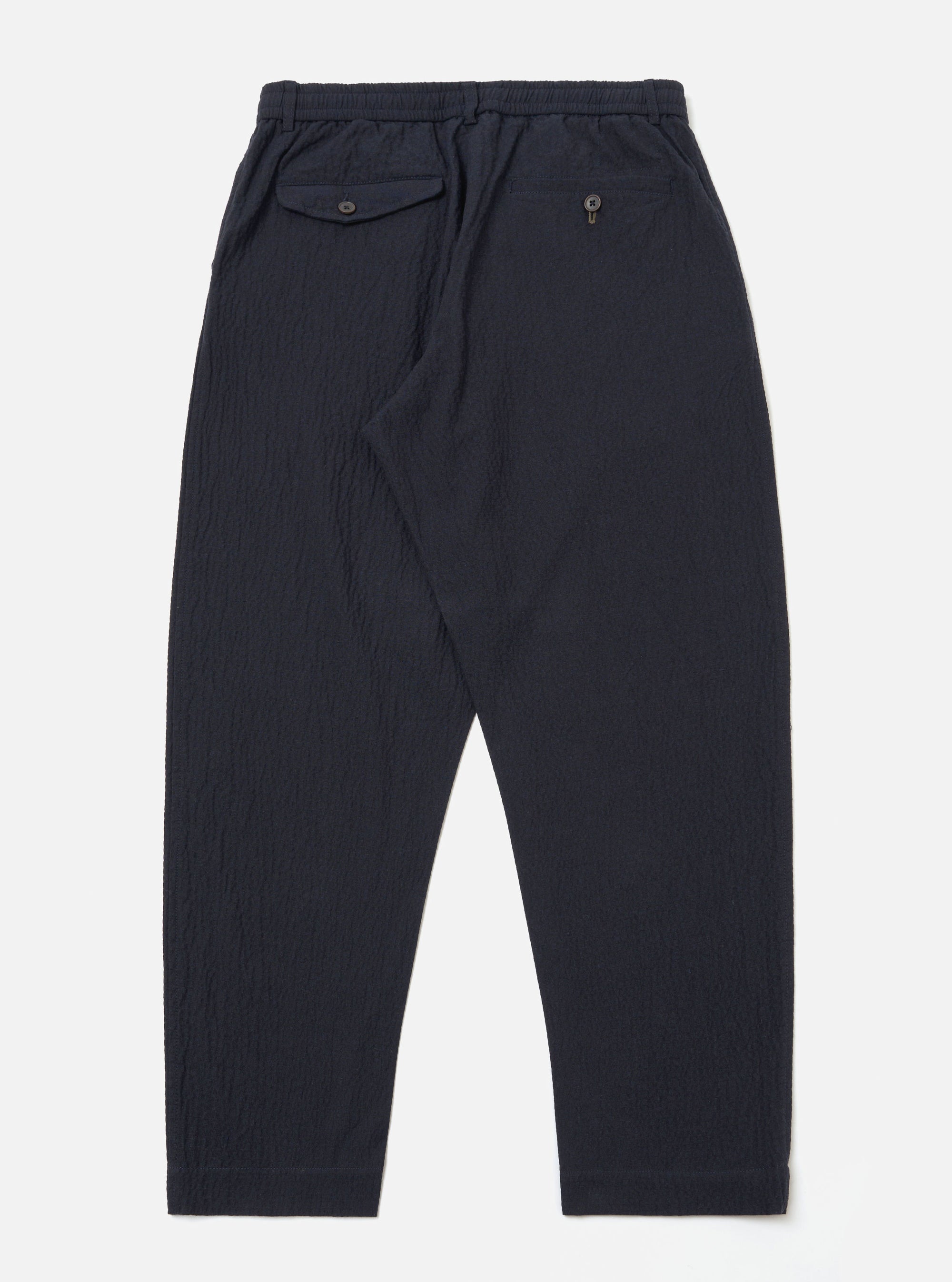 The Dark Navy Ospina Cotton Oxford Pant is shown from the back as a flat lay against a plain white background. The image highlights the elasticated waistband with a drawstring and two rear pockets: one with a buttoned flap and one with a buttoned jet. The dark navy fabric features a visible puckered seersucker texture.
