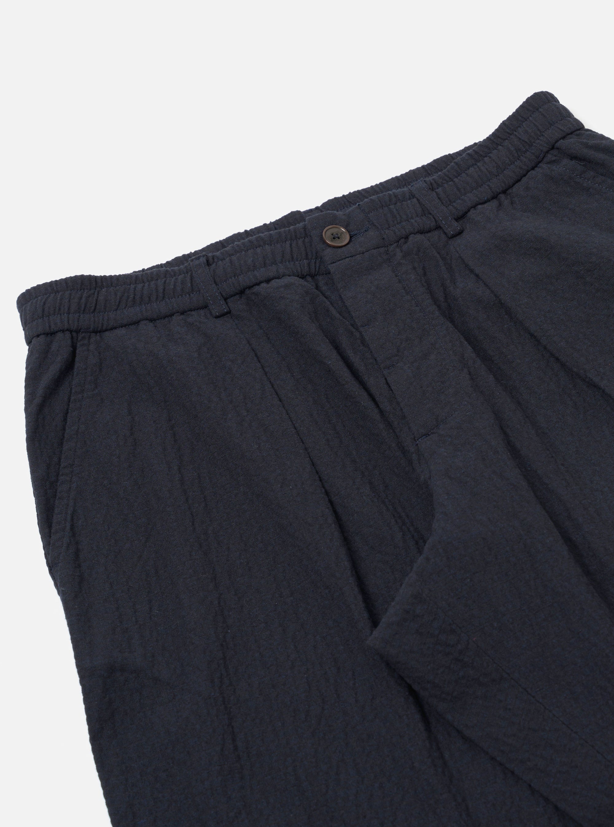 The Dark Navy Ospina Cotton Oxford Pant is shown from the back as a flat lay against a plain white background. The image highlights the two rear pockets, one with a buttoned flap and one with a buttoned jet, as well as the elasticated waistband. The dark navy fabric features a subtle, puckered seersucker texture.