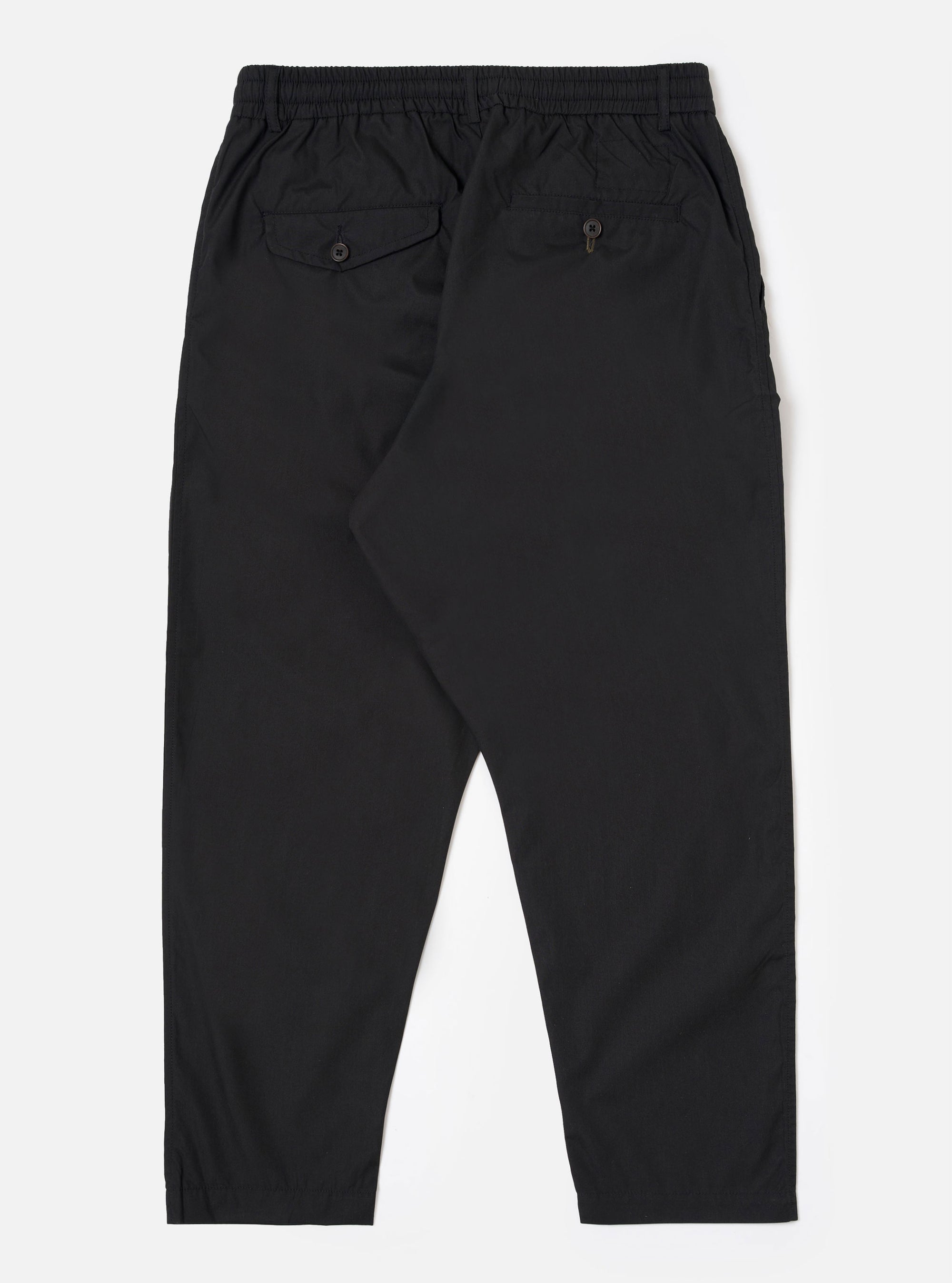 The Black Recycled Polytech Pleated Track Pant is shown from the rear as a flat lay against a plain white background. The image highlights the elasticated waistband and two rear pockets, including one with a buttoned flap on the left and a buttoned jetted pocket on the right. The black fabric has a smooth, technical appearance.