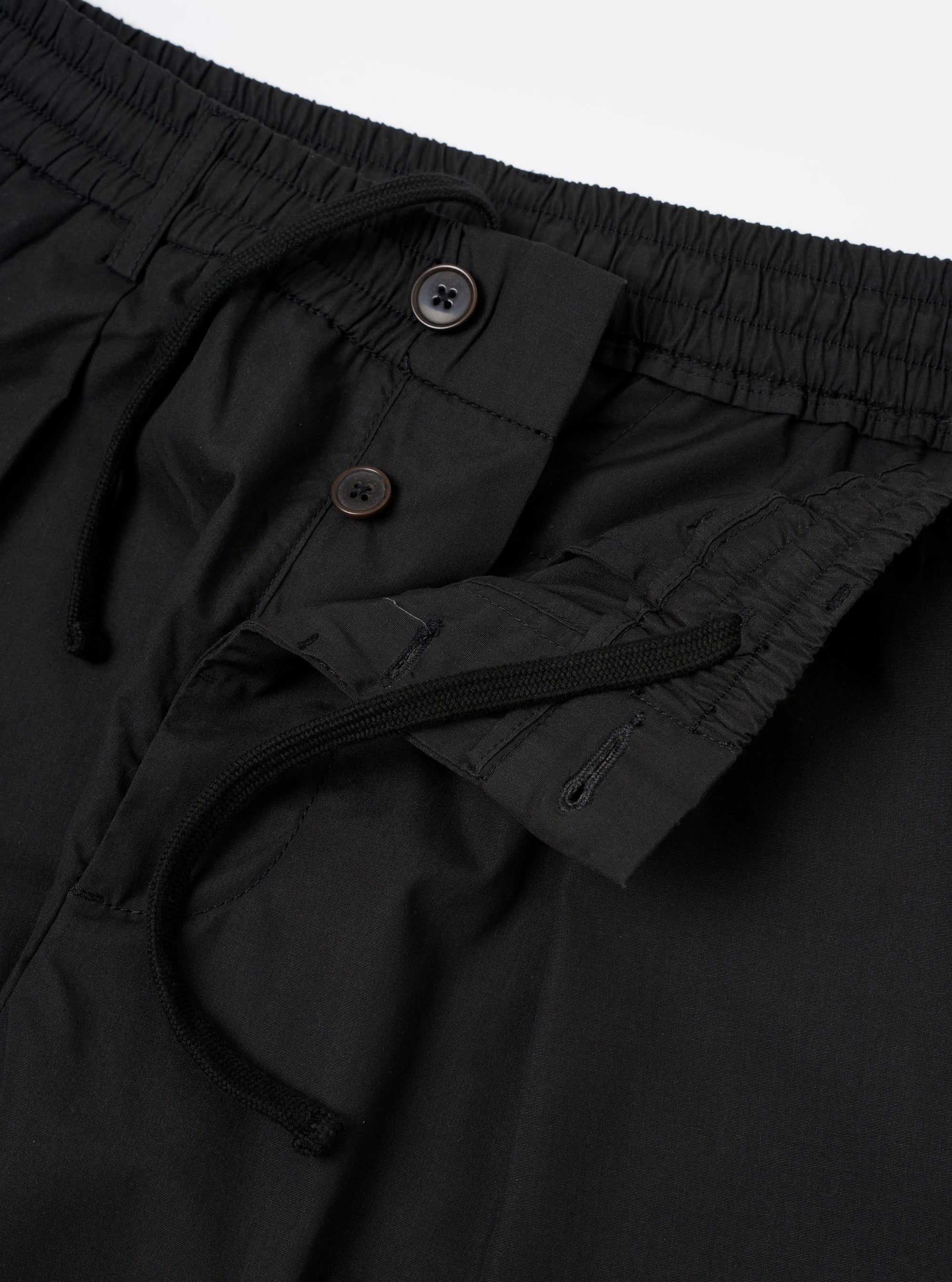 The Black Recycled Polytech Pleated Track Pant is shown from a rear three-quarter angle as a flat lay against a plain white background. The image highlights the tapered leg, a rear patch pocket with a buttoned flap, and the smooth, technical texture of the black recycled polytech fabric.