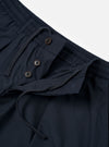 The Navy Recycled Polytech Pleated Track Pant is shown from the rear three-quarter angle as a flat lay against a plain white background. The image highlights the relaxed, tapered fit of the legs, the elasticated waistband, and the two rear pockets, including one with a button-down flap. The dark navy fabric has a smooth, technical finish.