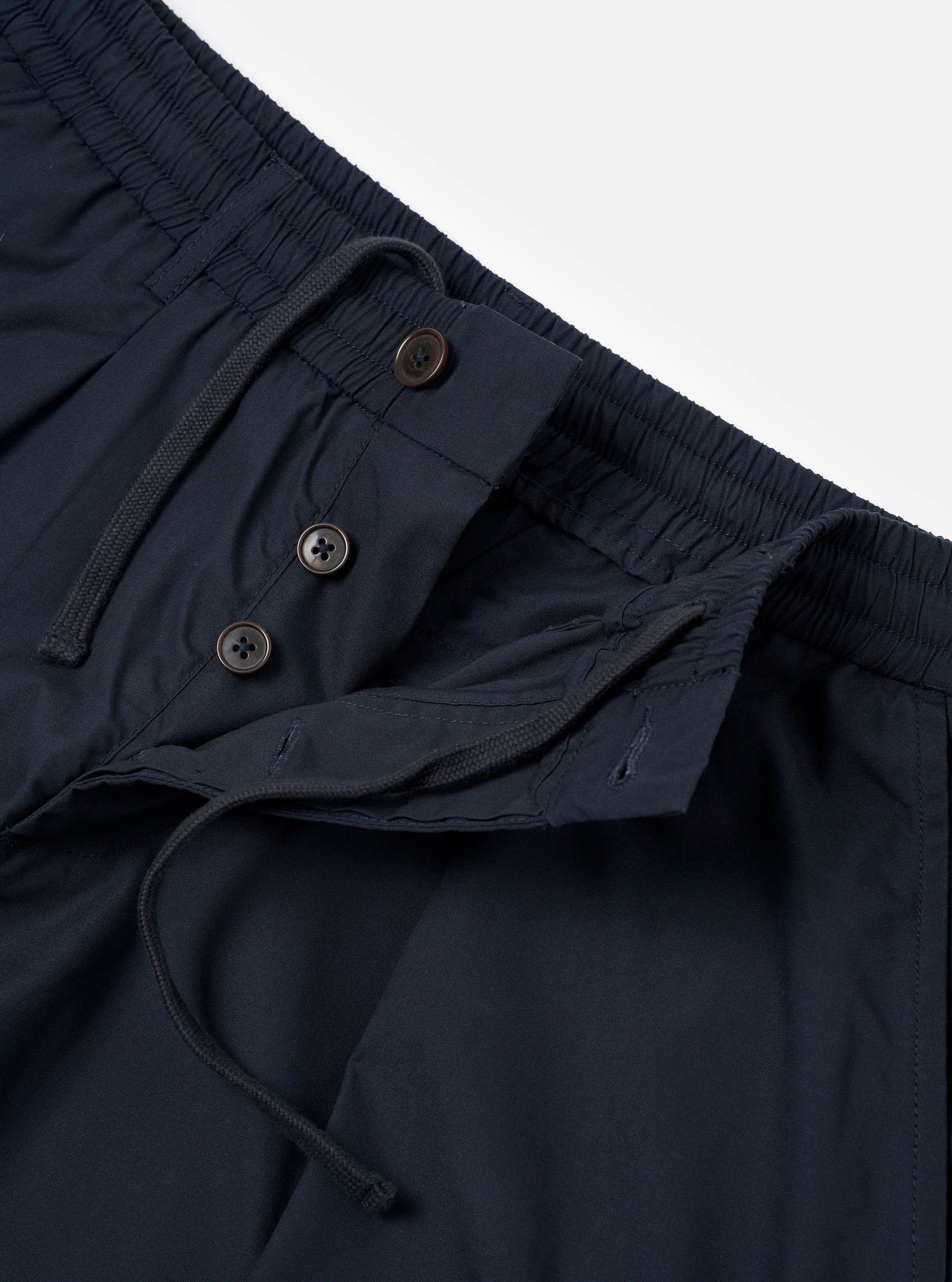 The Navy Recycled Polytech Pleated Track Pant is shown from the rear three-quarter angle as a flat lay against a plain white background. The image highlights the relaxed, tapered fit of the legs, the elasticated waistband, and the two rear pockets, including one with a button-down flap. The dark navy fabric has a smooth, technical finish.