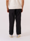 A model wears the Black Recycled Polytech Pleated Track Pant, shown from the back against a plain white background. The image highlights the relaxed, tapered fit of the trousers and the clean, utilitarian appearance of the smooth black recycled polytech fabric.