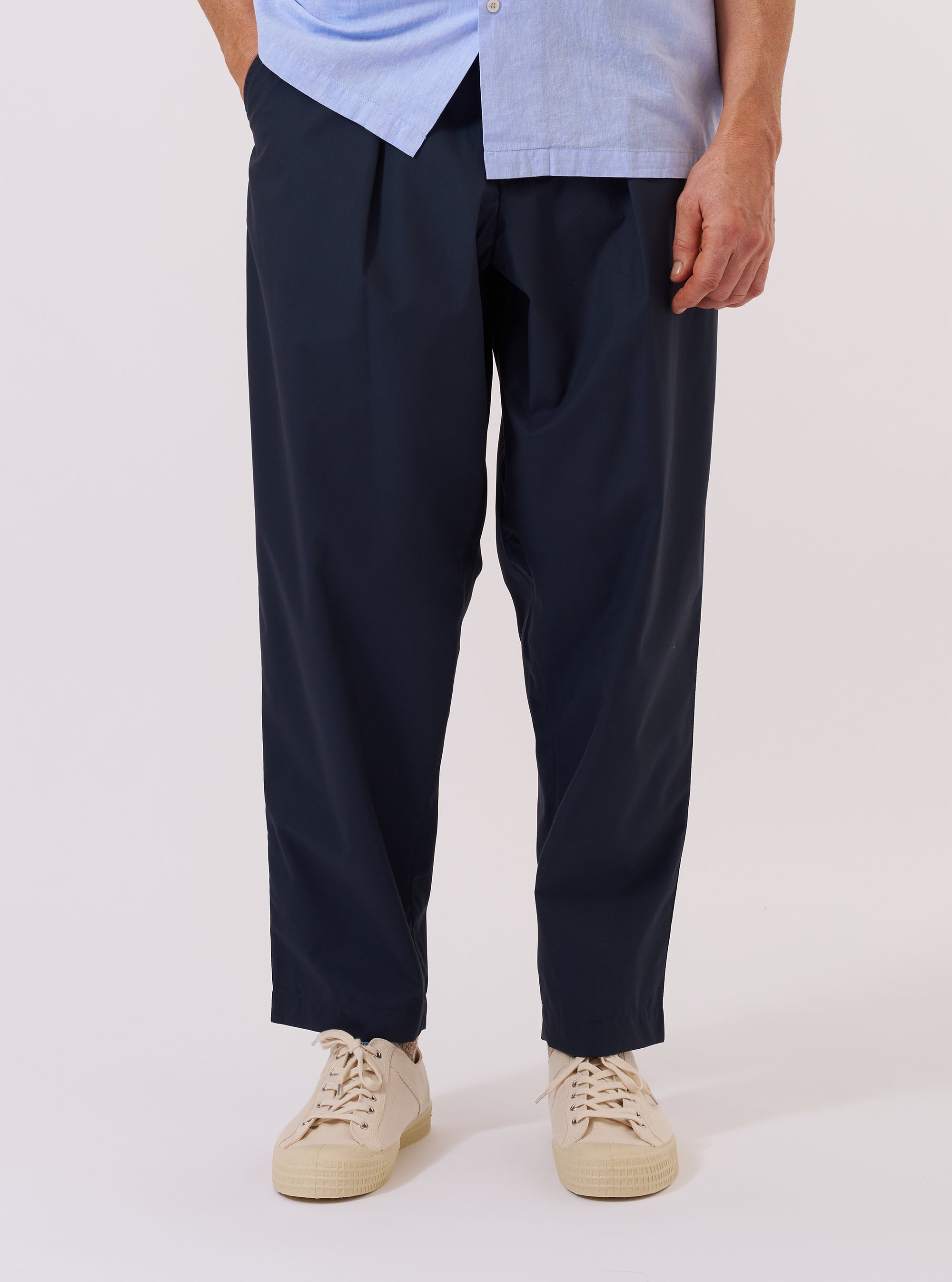 A model wears the Navy Recycled Polytech Pleated Track Pant while standing against a plain, neutral studio background. The image shows a front view of the trousers, highlighting the loose fit, tapered leg, and single pleat detail. The model is wearing the trousers with a light-colored shirt tucked in, showcasing the elasticated waistband and internal drawstring. The navy blue recycled polytech fabric has a smooth, utilitarian finish. No mannequin is present in the image.
