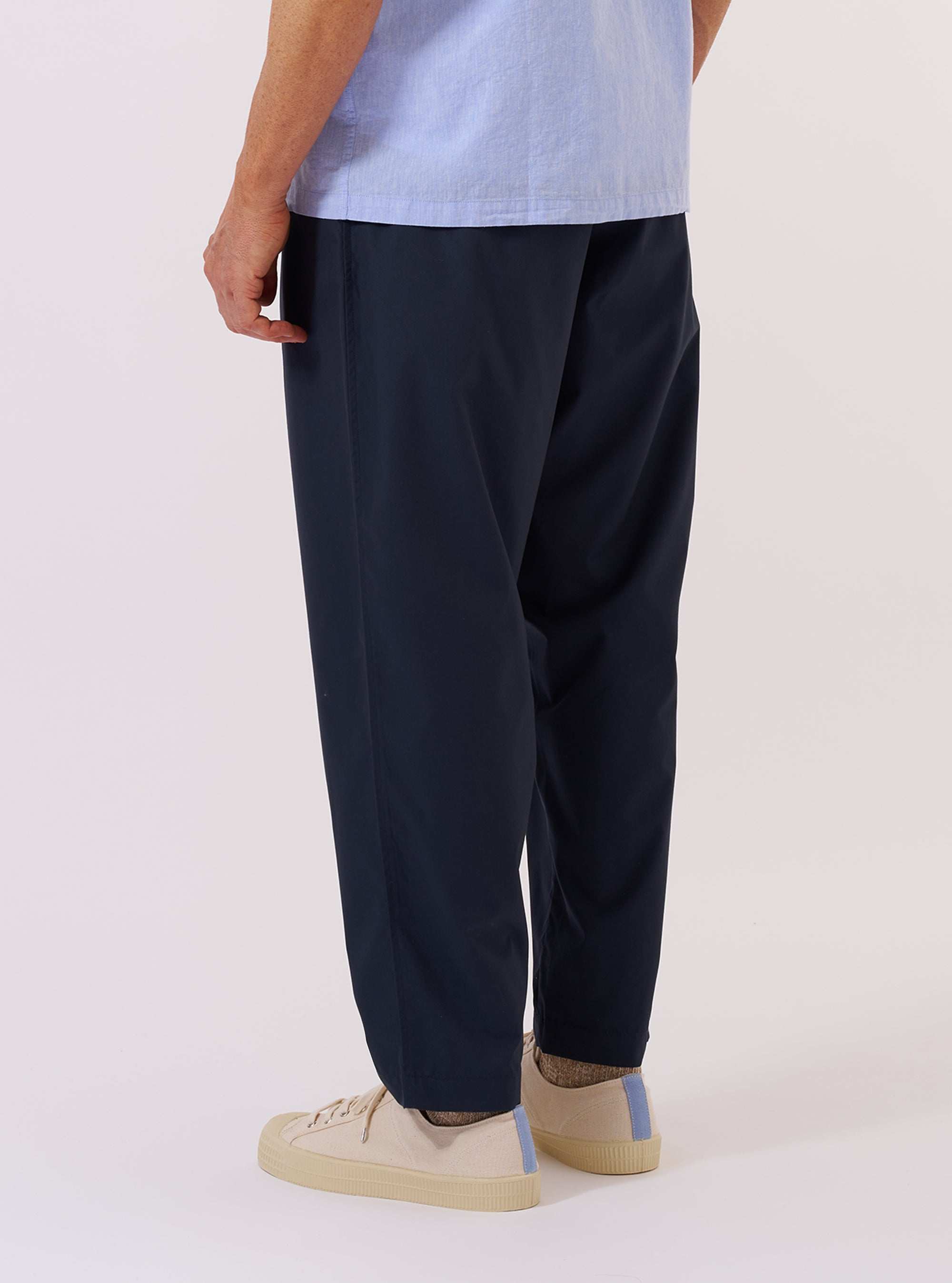 A model wears the Navy Recycled Polytech Pleated Track Pant, shown from the waist down against a plain white background. The image highlights the loose silhouette, the single pleat at the front, and the tapered leg. The model is also wearing white socks and dark shoes.
