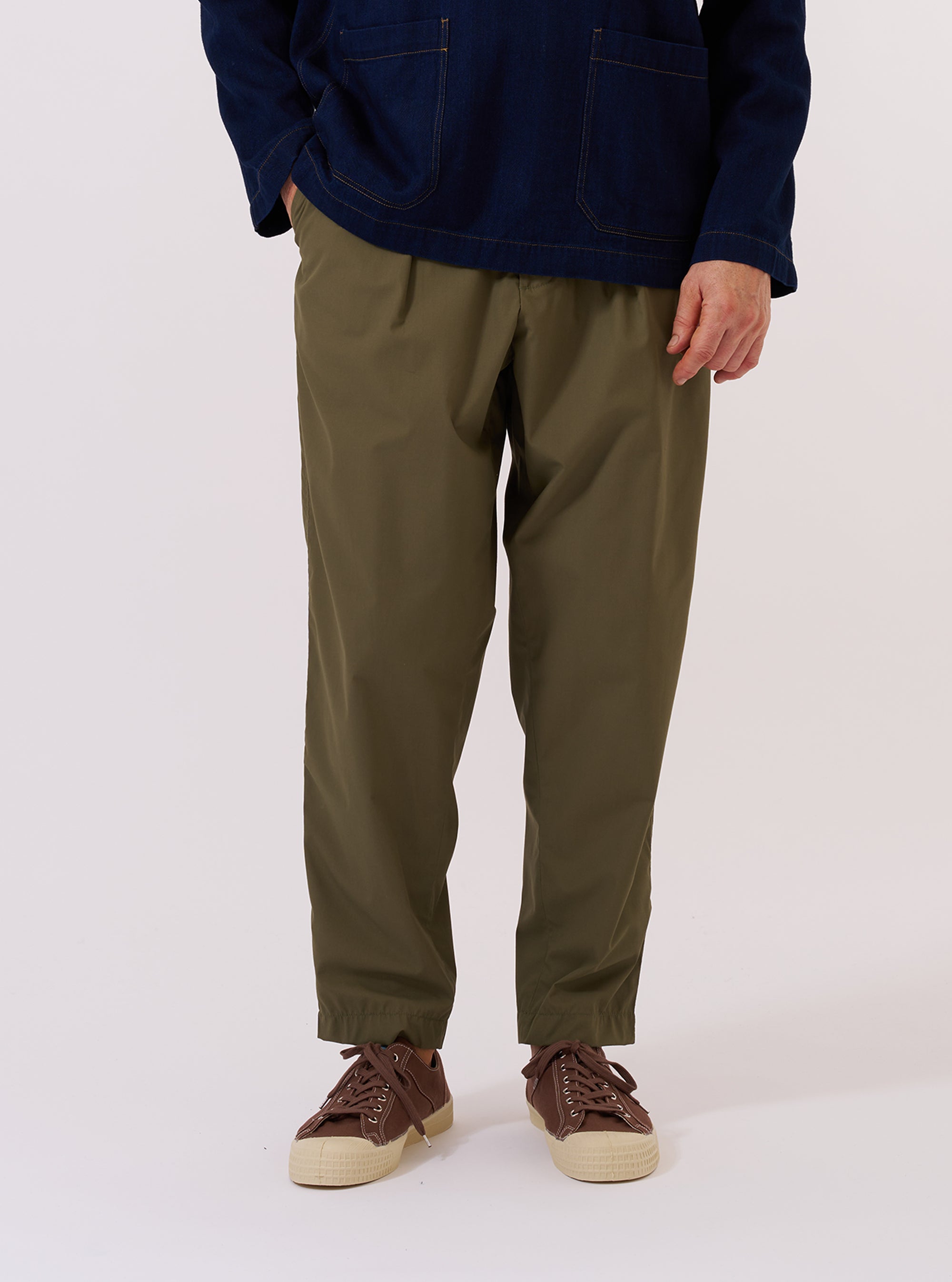 A model wears the Olive Recycled Polytech Pleated Track Pant while standing against a plain white background. The trousers feature a loose fit with a tapered leg, a single pleat at the front, and an elasticated waistband with a drawstring. The model is wearing the pants with a white t-shirt and white socks, highlighting the smooth, olive green technical fabric.