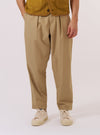 A model wears the Sand Recycled Polytech Pleated Track Pant while standing against a plain white background. The image shows a front view of the trousers, highlighting the loose fit, tapered leg, and the single pleat at the front. The elasticated waistband and the light sand-colored recycled poly-tech fabric are prominent. The model is wearing the trousers with a light-colored top tucked in to display the waistband detail.