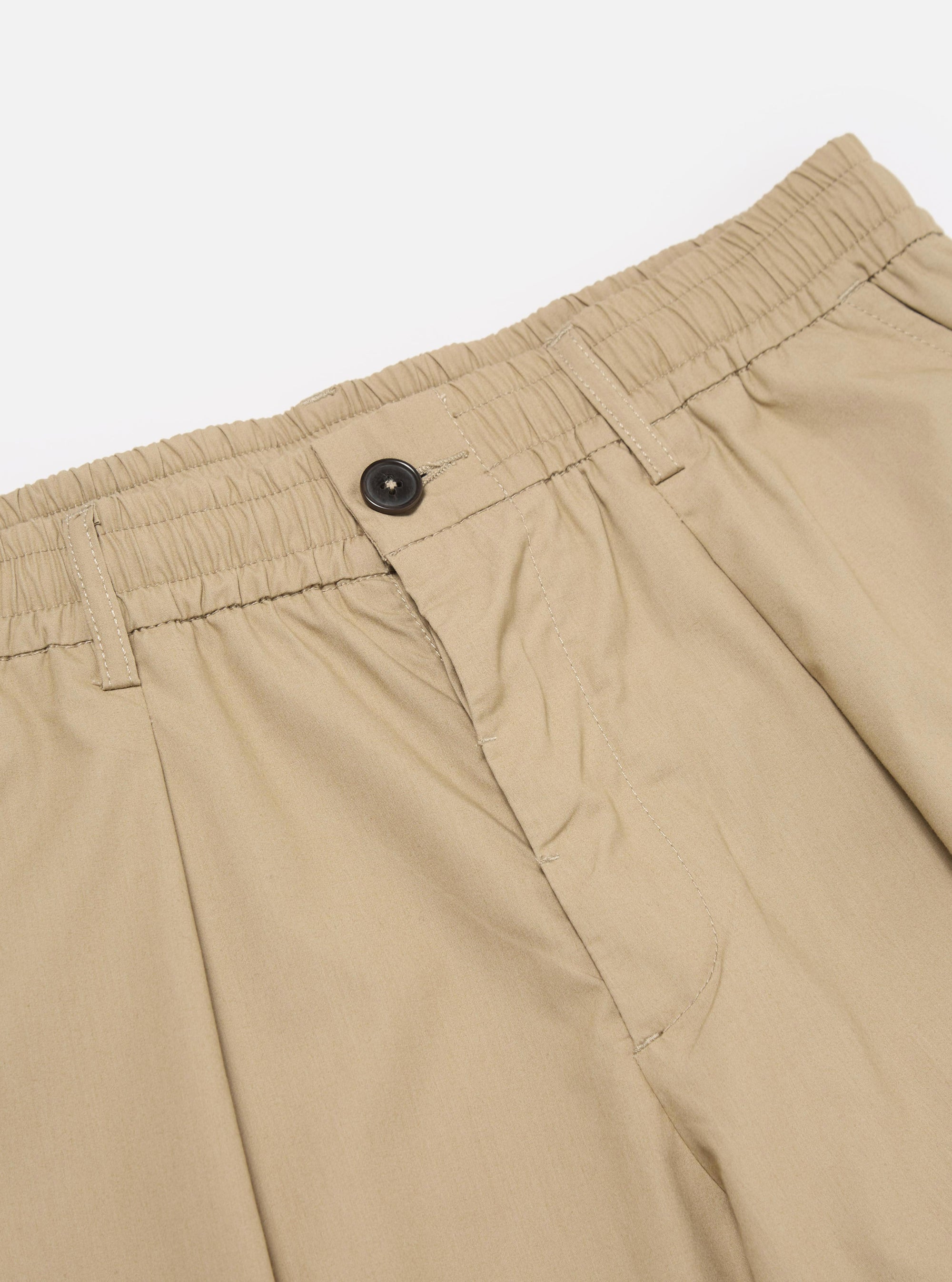 The Sand Recycled Polytech Pleated Track Pant is shown from the back as a flat lay against a plain white background. The image highlights the elasticated waistband and the two rear pockets, including one with a buttoned flap and one jetted pocket with a button closure. The fabric is a smooth, sand-colored technical blend.