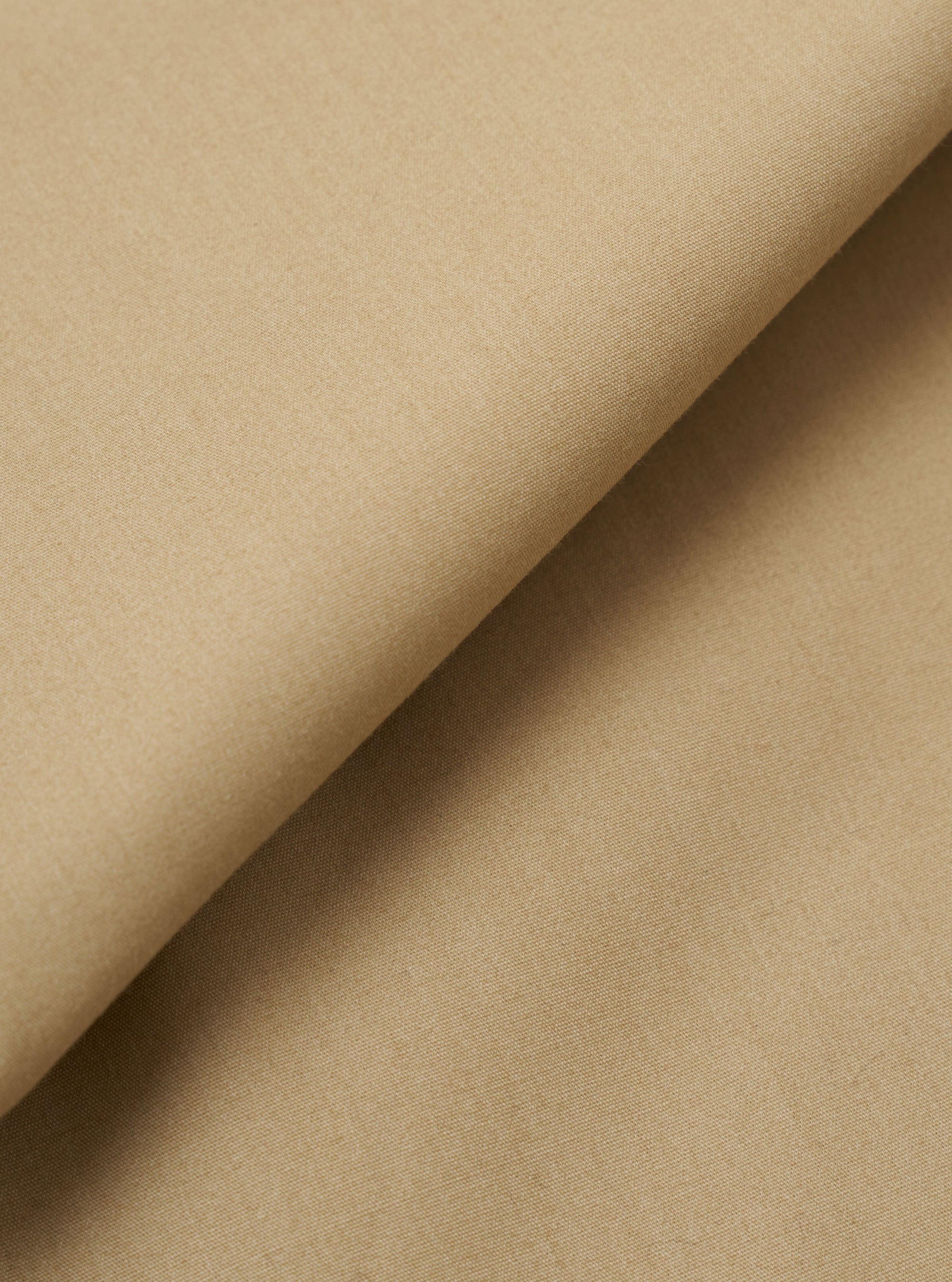 A close-up fabric swatch of the Sand Recycled Polytech Pleated Track Pant is shown, highlighting the light sand color and the smooth, technical weave of the recycled polyester and cotton blend.