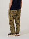 A model wears the Sand Jungle Toile Twill Oxford Pant, shown from the waist down against a plain white background. The pants feature a loose, wide-leg fit and an intricate dark jungle toile print on a sand-colored base. The model is standing and wearing the pants with white socks and dark shoes, highlighting the pleated front and relaxed drape of the fabric.