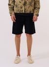 A model wears the Black Organic Fine Cord Fatigue Short while standing against a plain white background. The image shows the front view of the shorts, highlighting the two large military-inspired patch pockets, a zip fly, and belt loops. The garment is made from a fine, narrow-wale black corduroy fabric and features a relaxed fit that ends just above the knee.