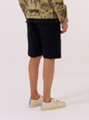 A model is wearing the Black Organic Fine Cord Fatigue Short, shown from the waist down against a plain white background. The image captures a rear three-quarter view, highlighting the relaxed fit, the fine corduroy texture of the black organic cotton, and a large rear patch pocket. The model is wearing a light-colored long-sleeve shirt tucked into the shorts.