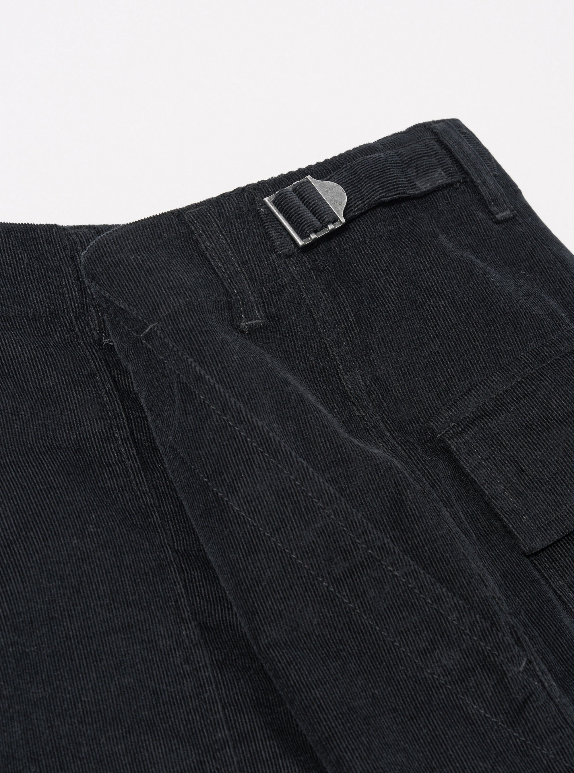 The Black Organic Fine Cord Fatigue Short is shown from the back as a flat lay against a plain white background. The image highlights the two rear patch pockets, one of which features a button-through closure, and the elasticated waistband. The black organic cotton fabric has a visible fine-wale corduroy texture.