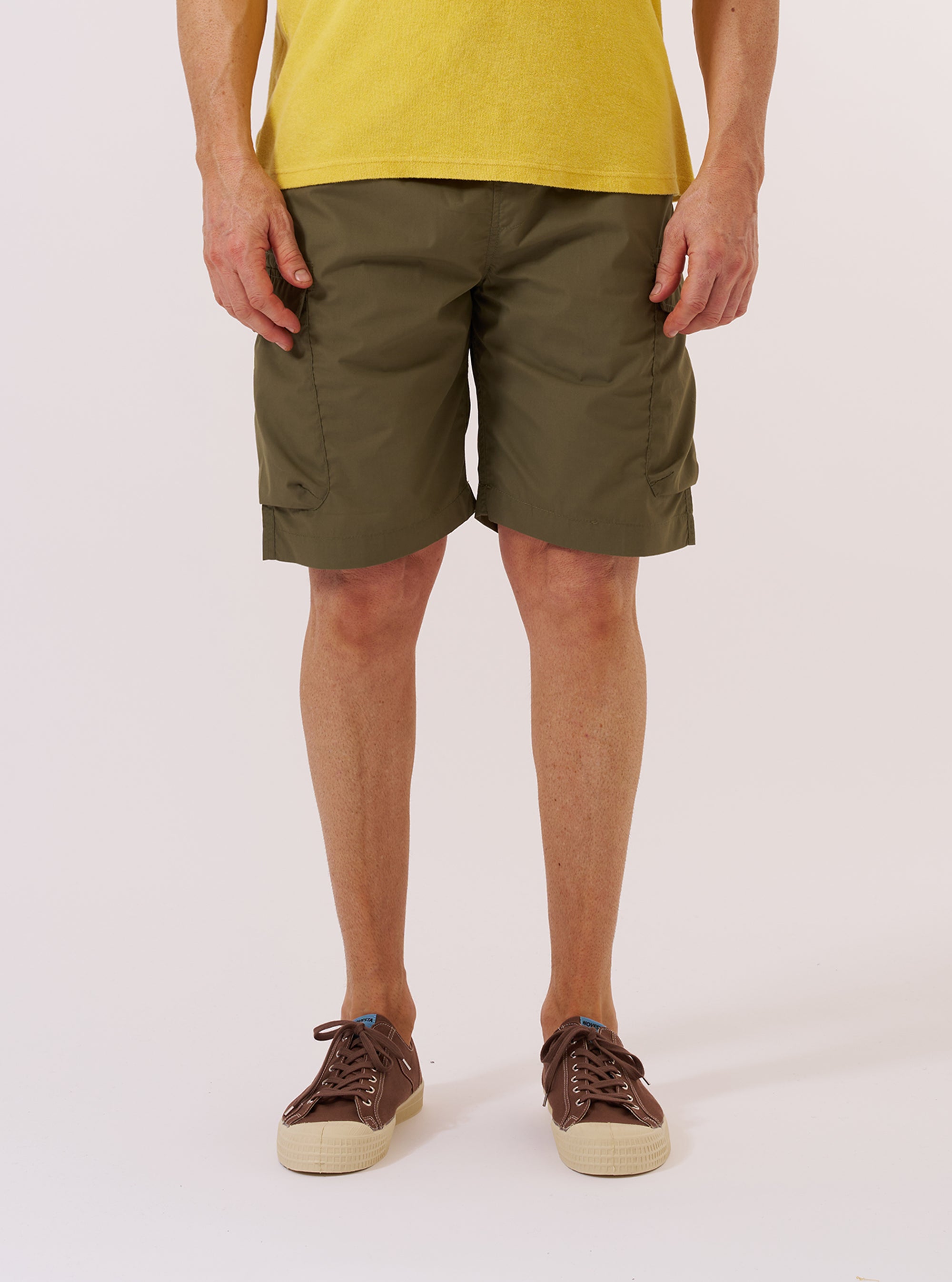 A model wears the Olive Recycled Polytech Parachute Short while standing against a plain white background. The shorts feature a relaxed, wide-leg fit, an elasticated waistband with an integrated drawstring, and large side pockets. The garment is made from a smooth, olive green technical fabric. The model is shown from the waist down, providing a clear view of the front and side profile of the shorts.