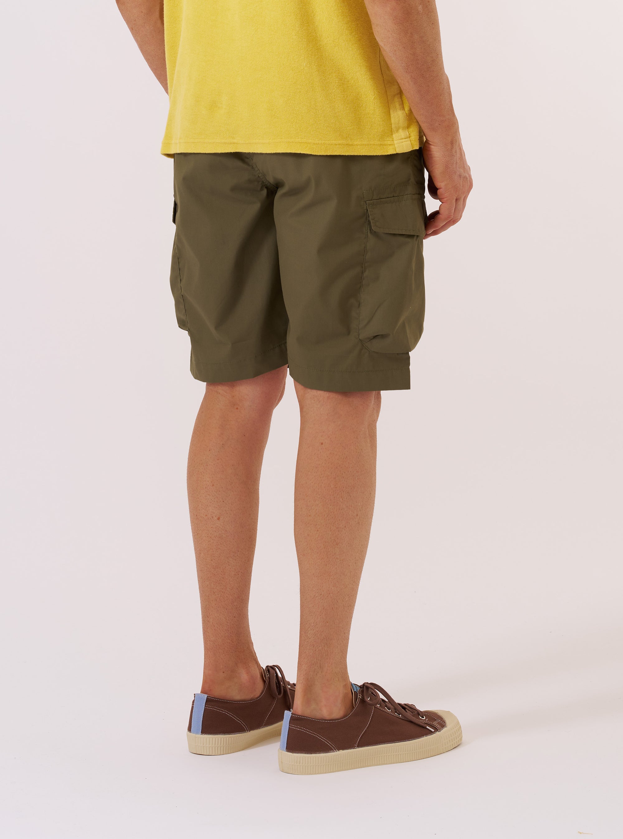 The Olive Recycled Polytech Parachute Short is worn by a model against a plain white background. The image shows the shorts from the back, highlighting the two rear patch pockets and the relaxed, knee-length fit. The model wears the shorts with a light-colored shirt and dark shoes.