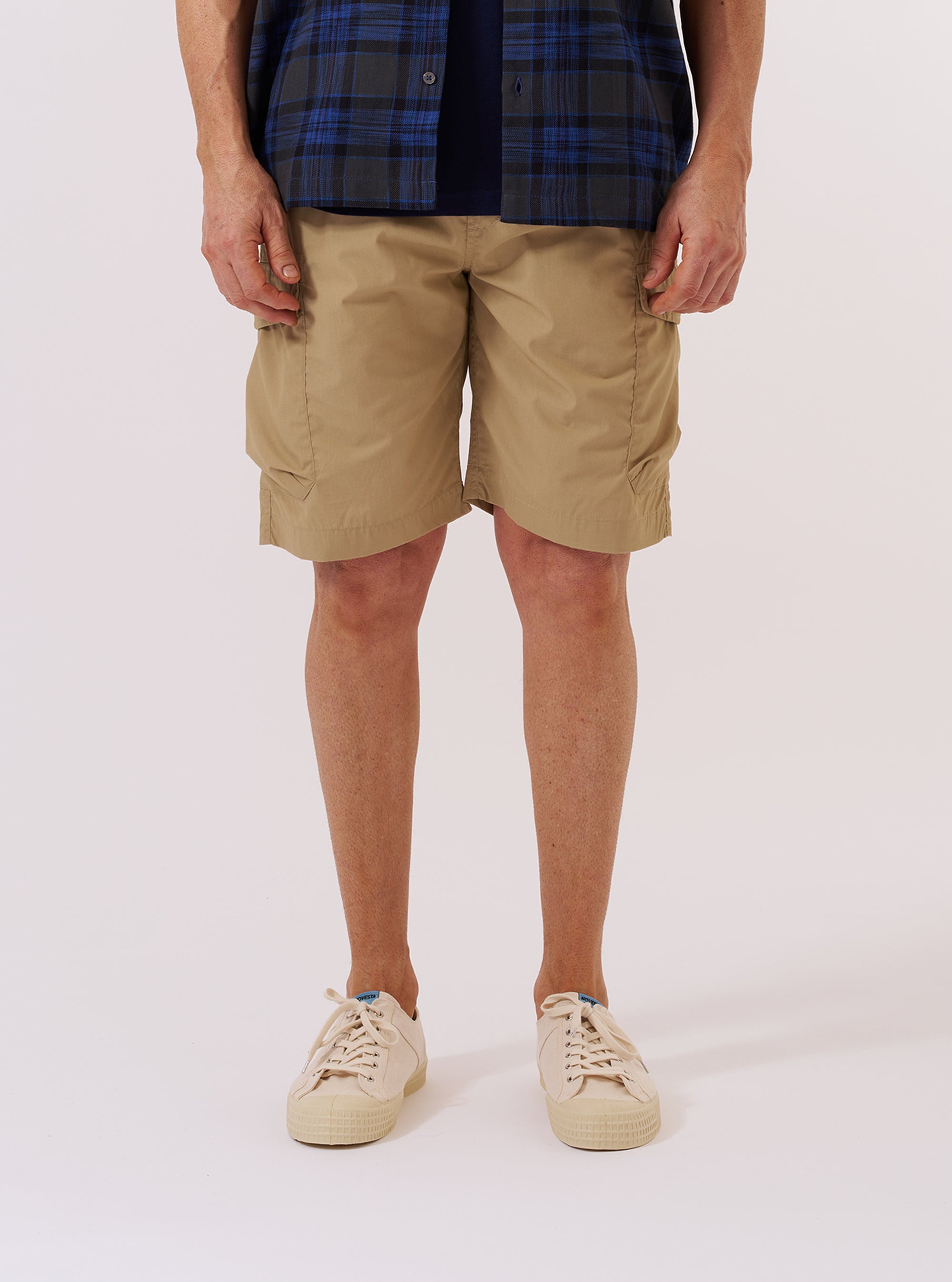 A model wears the Sand Recycled Polytech Parachute Short over a white long-sleeved shirt against a plain white background. The image shows a front view of the shorts, highlighting the relaxed fit, knee-length cut, and large military-style bellows pockets on the sides. The elasticated waistband with a drawstring is also visible.
